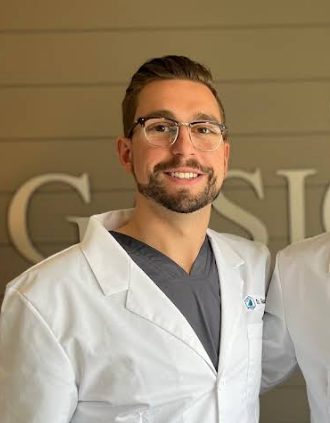Dr. Max Gosselin, a dentist at Gasior Family Dental, wearing glasses and a white lab coat over gray scrubs, standing indoors against a neutral wall with partial lettering visible in the background.