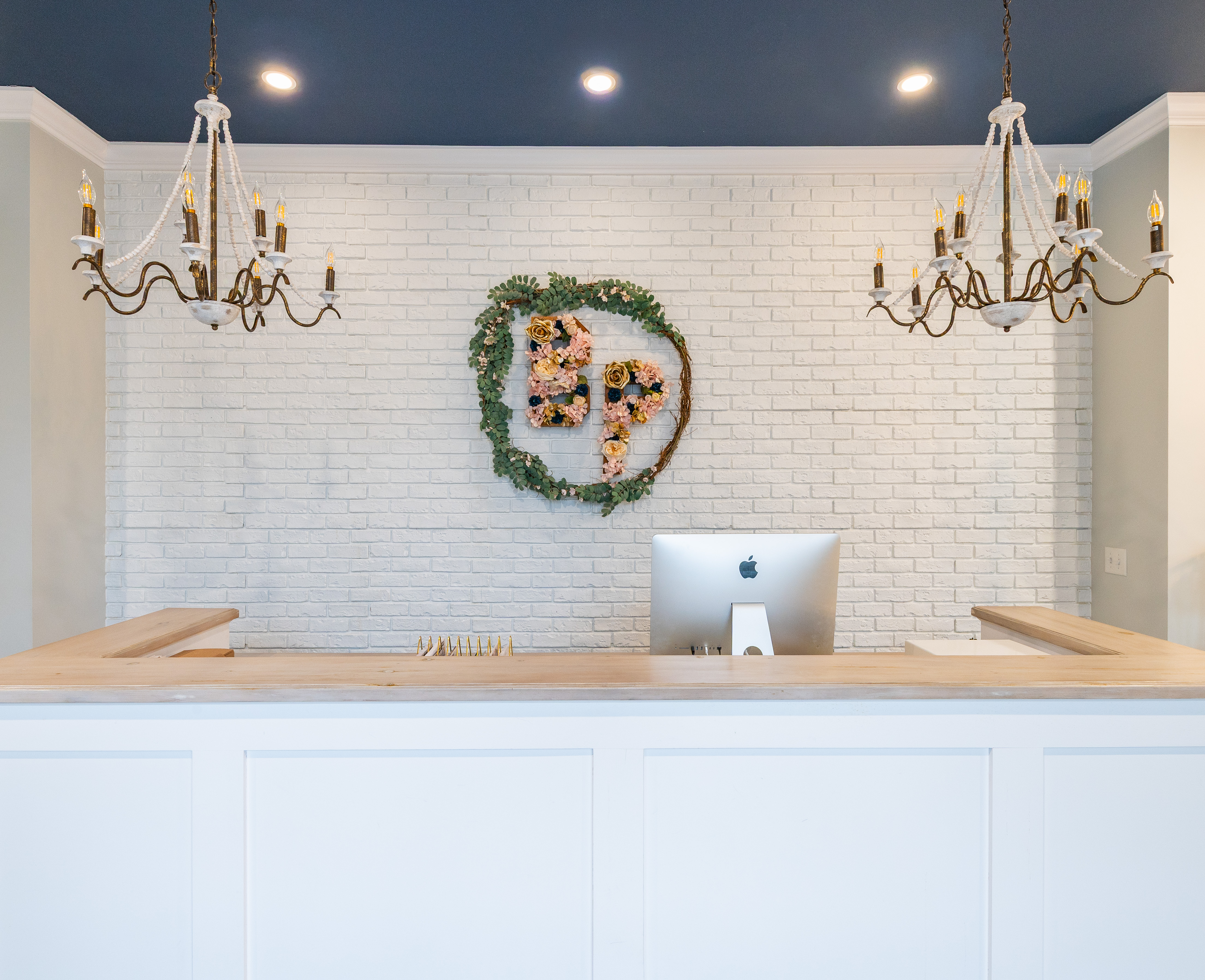 Minimalist reception desk with wood countertop, two chandeliers, and a floral wreath logo on a white brick wall.