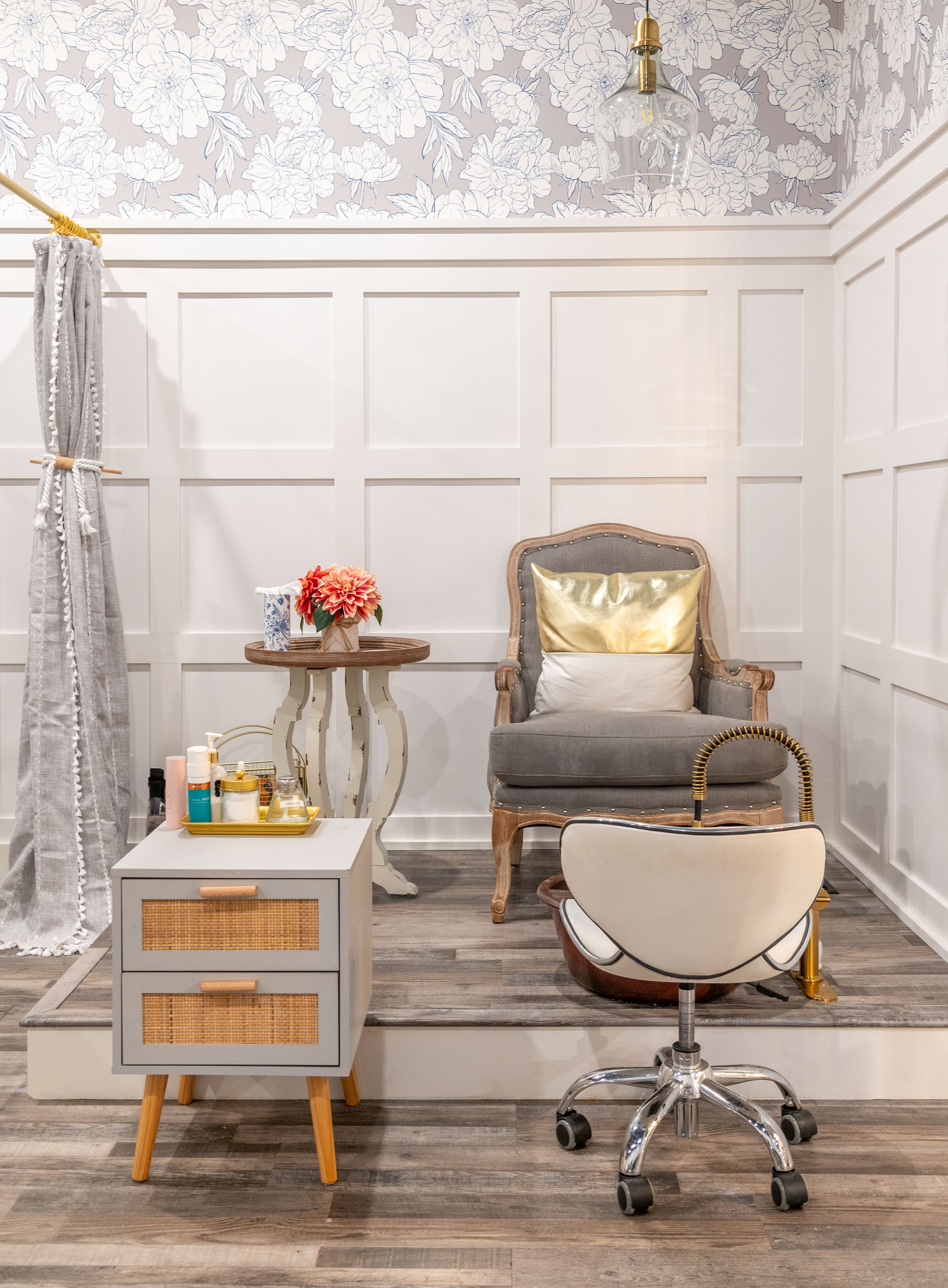 Cozy nail salon corner with wooden armchair with gold pillow, rolling white stool, floral wallpaper, and small side table with flower arrangement.