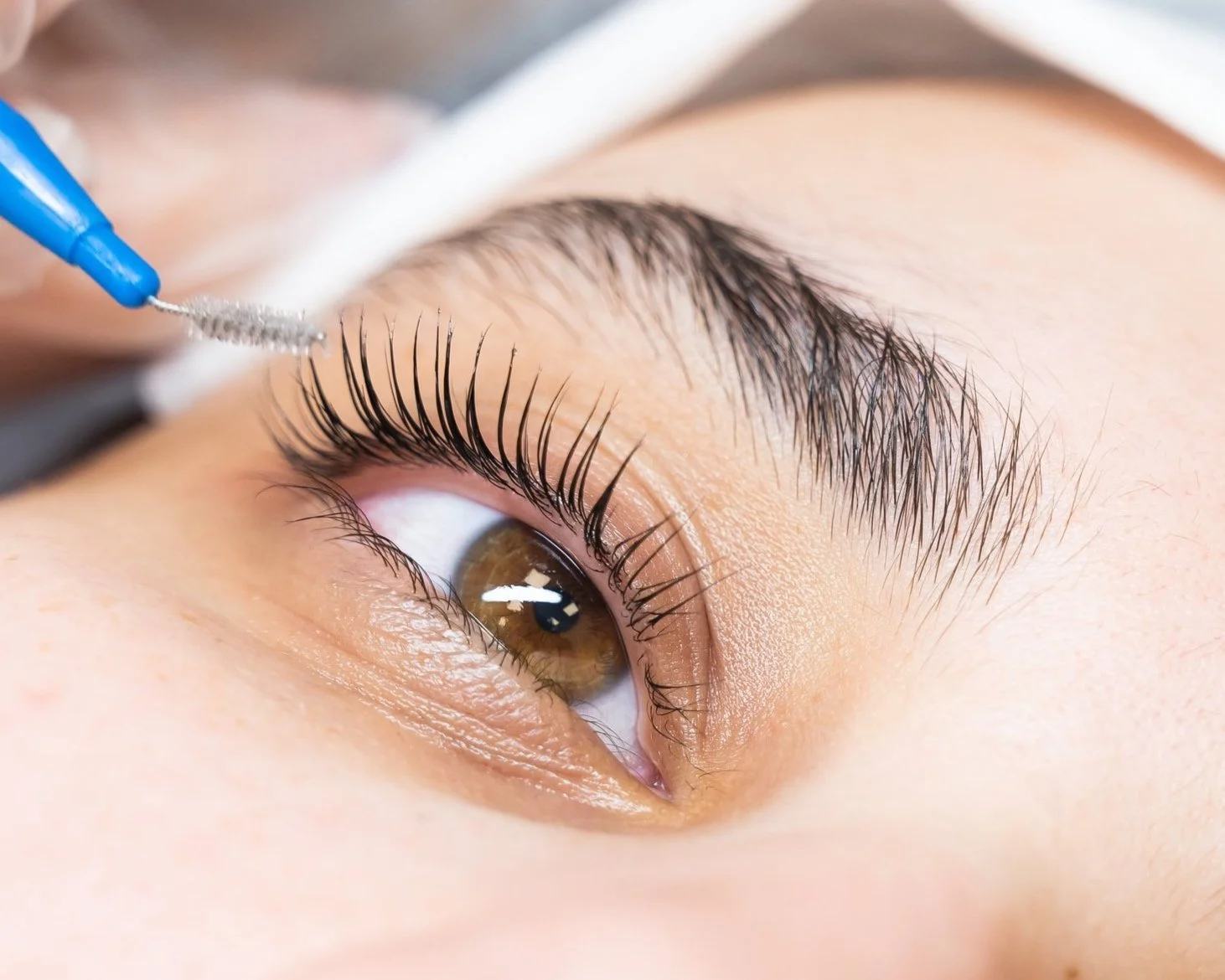 Close-up of an eye with brown iris and long eyelashes being brushed with a spoolie applicator.