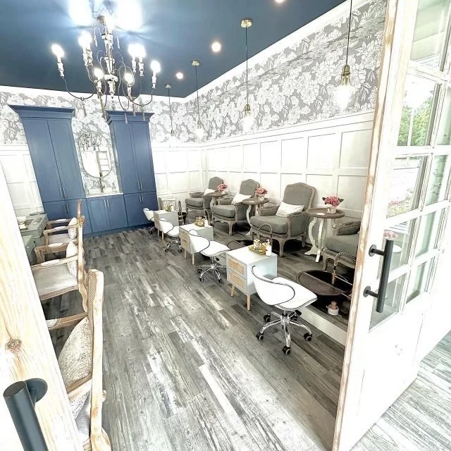 Elegant nail salon interior with white swivel chairs, gray armchairs, floral wallpaper, blue cabinetry, and wooden flooring.