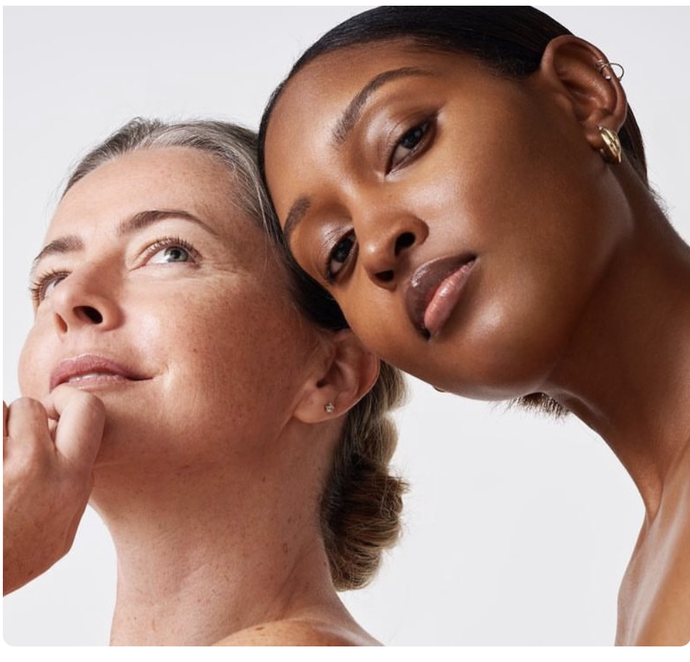 Close-up of two women with clear skin, one with light skin and gray hair looking thoughtfully upward, and the other with dark skin and dark hair leaning her head against the first woman.