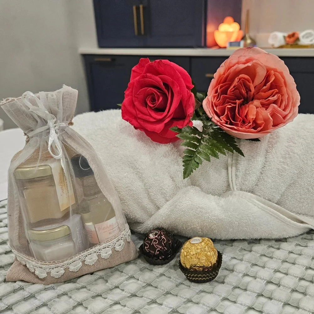 Spa setup with a white towel adorned with a red and a peach rose, a small bag of spa products, and two chocolates on a textured surface.
