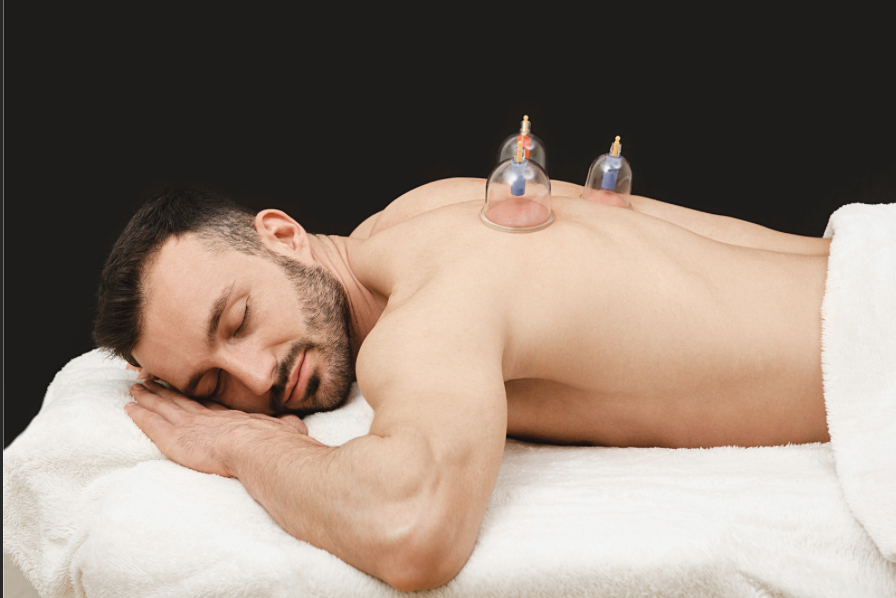 Man lying face down on a white towel with two cupping therapy cups on his back against a black background.
