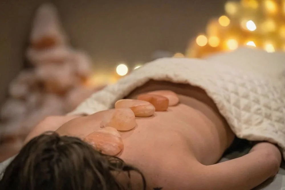 Person lying face down with large smooth stones on their back under a blanket in a relaxing spa setting.