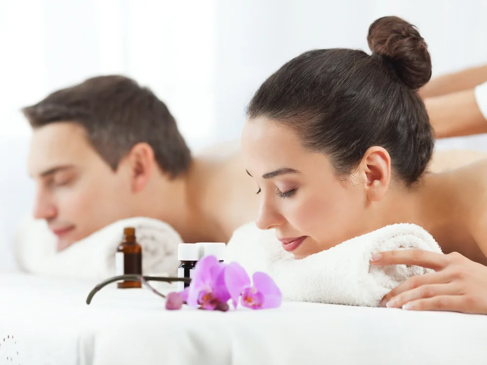 Woman and man receiving aromatherapy massages lying face down with closed eyes and towels under their heads, surrounded by essential oil bottles and purple orchid flowers.