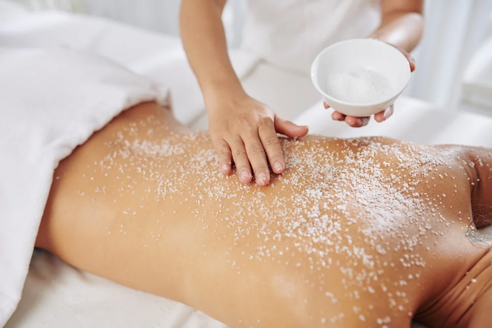 Person applying salt body scrub on another person's back during a spa treatment.