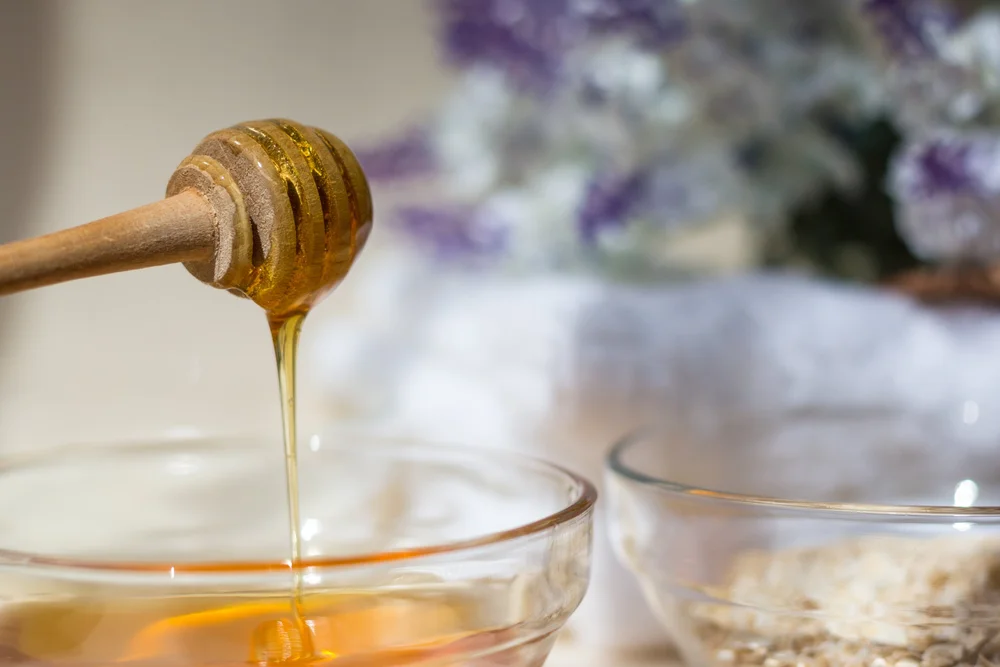 Honey dripping from a wooden honey dipper into a glass bowl with blurred lavender flowers in the background.