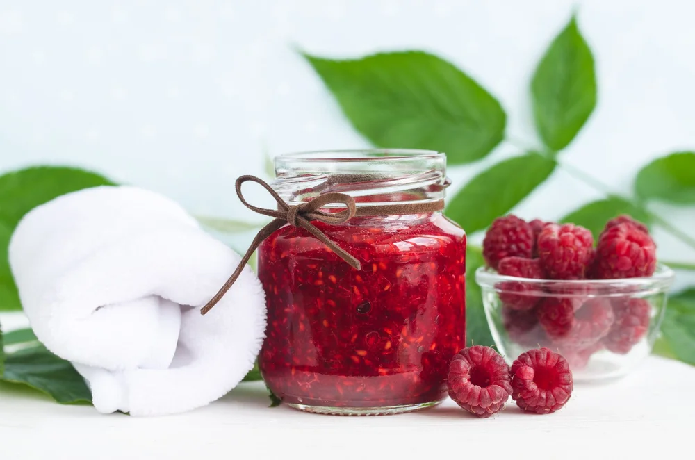 Jar of raspberry jam tied with a brown string, next to a rolled white towel and a bowl of fresh raspberries on a white surface with green leaves in the background.