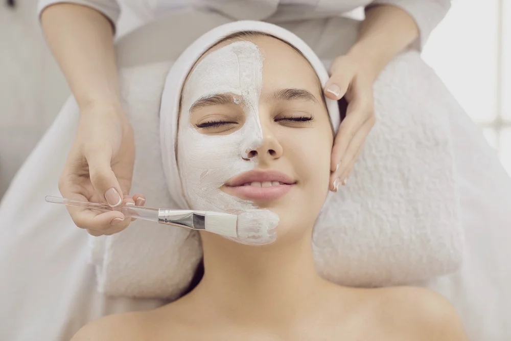 Woman with eyes closed receiving a white facial mask applied on half of her face at a spa treatment.