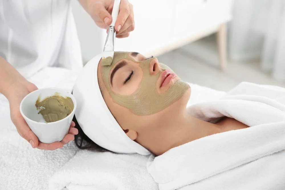 Woman with eyes closed receiving a green facial mask applied with a brush while lying on a spa bed.