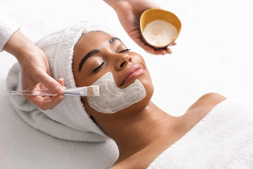 Woman with a towel wrapped around her head relaxing as a spa therapist applies a white facial mask with a brush to her cheek.