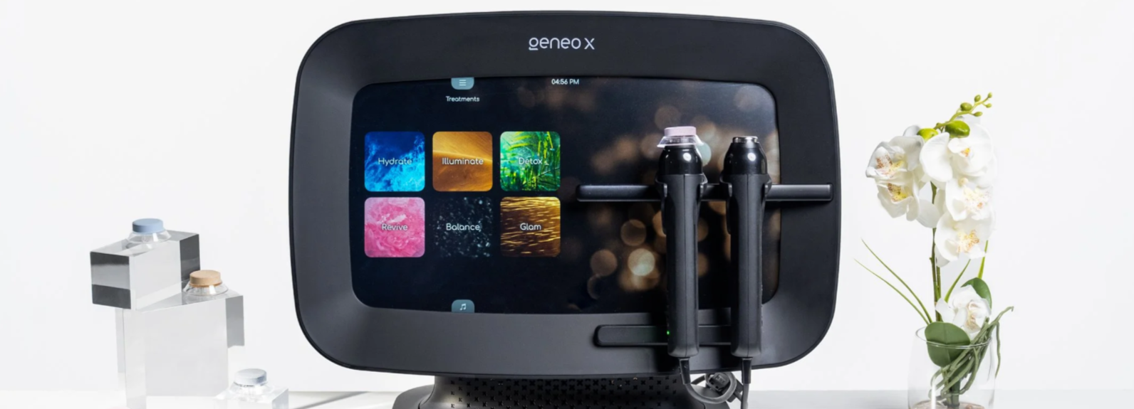 Geneo X facial treatment device with touchscreen displaying options Hydrate, Illuminate, Detox, Revive, Balance, and Glam, flanked by clear containers with caps on the left and white orchids in a glass vase on the right.