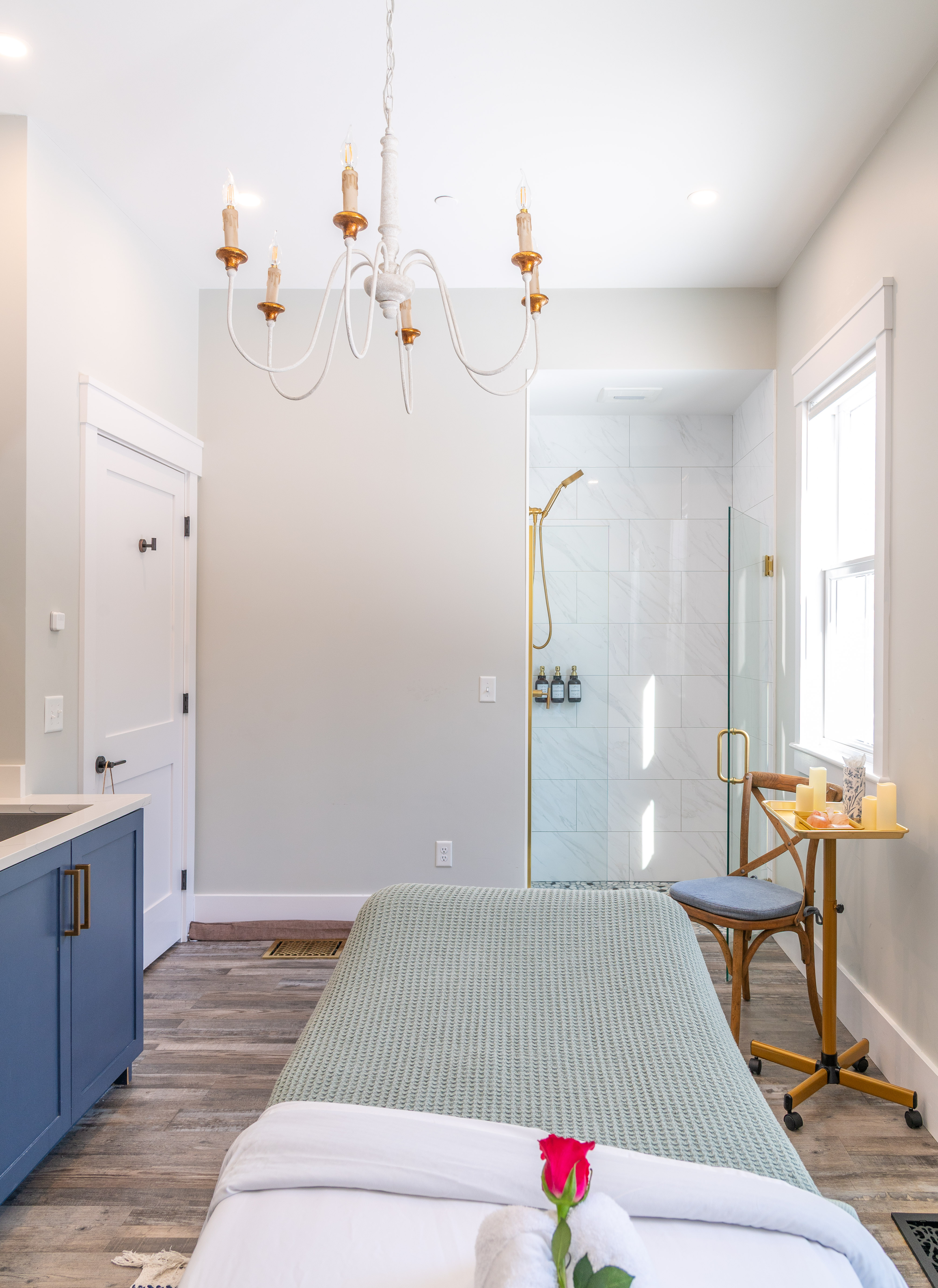 Bright massage room with blue cabinets, green bedcover, rose on towel, wooden chair, and glass shower.