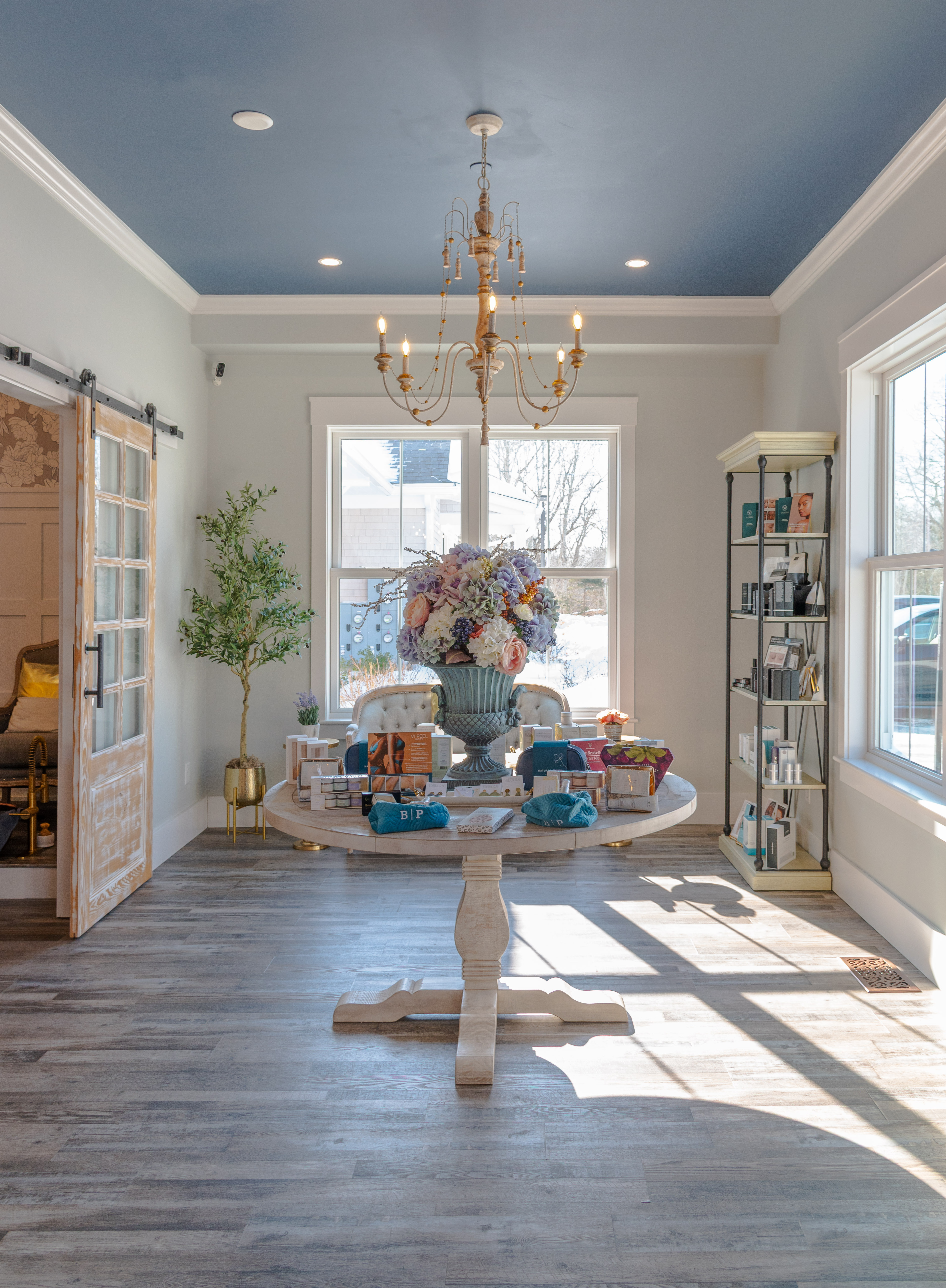 Bright room with a round wood table topped with floral arrangement and beauty products under chandelier.