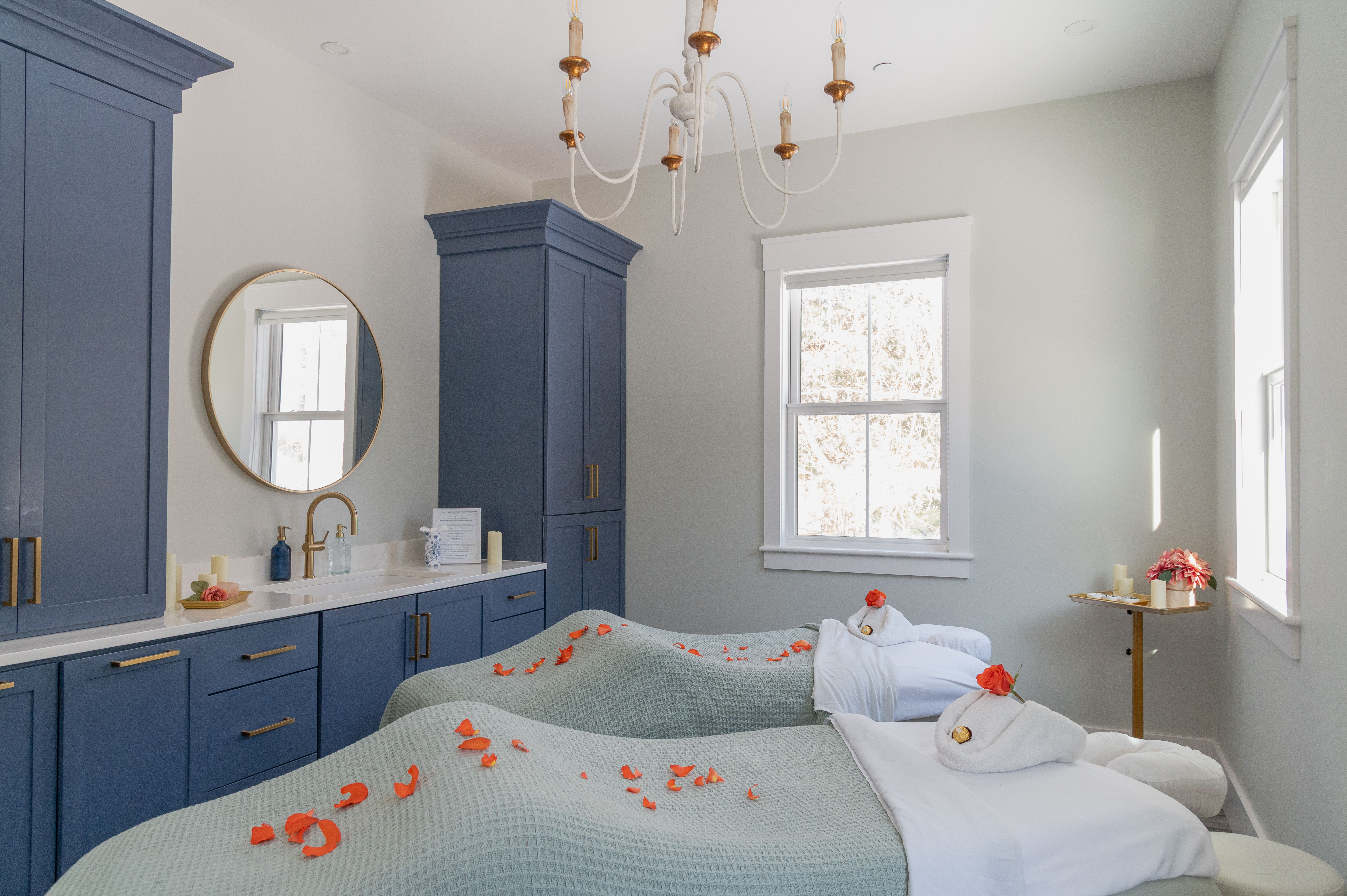 Bright spa room with two massage beds covered in green blankets and rose petals, blue cabinets, and a chandelier.