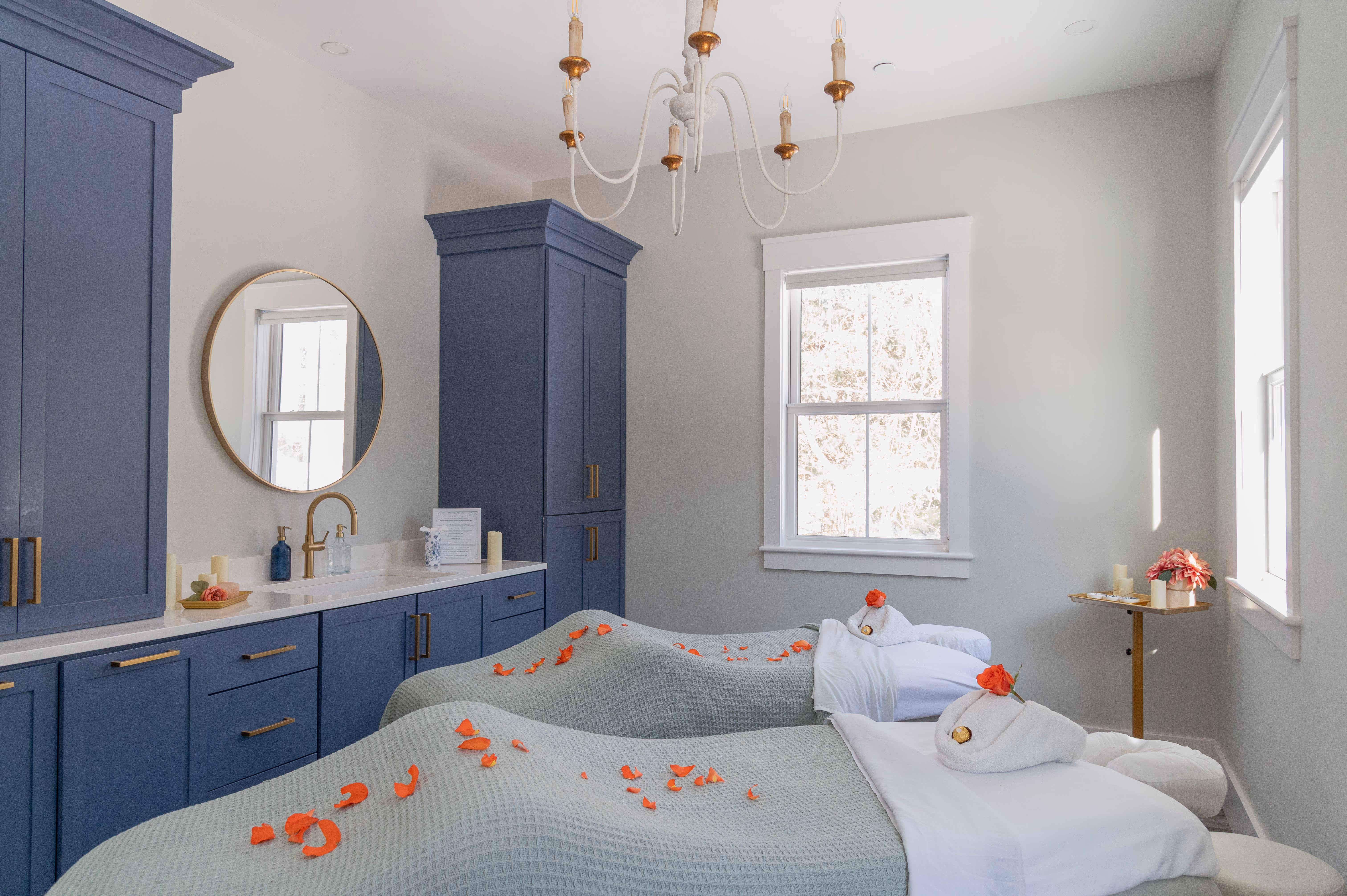 Bright spa room with two massage beds covered in green blankets and rose petals, blue cabinets, and a chandelier.