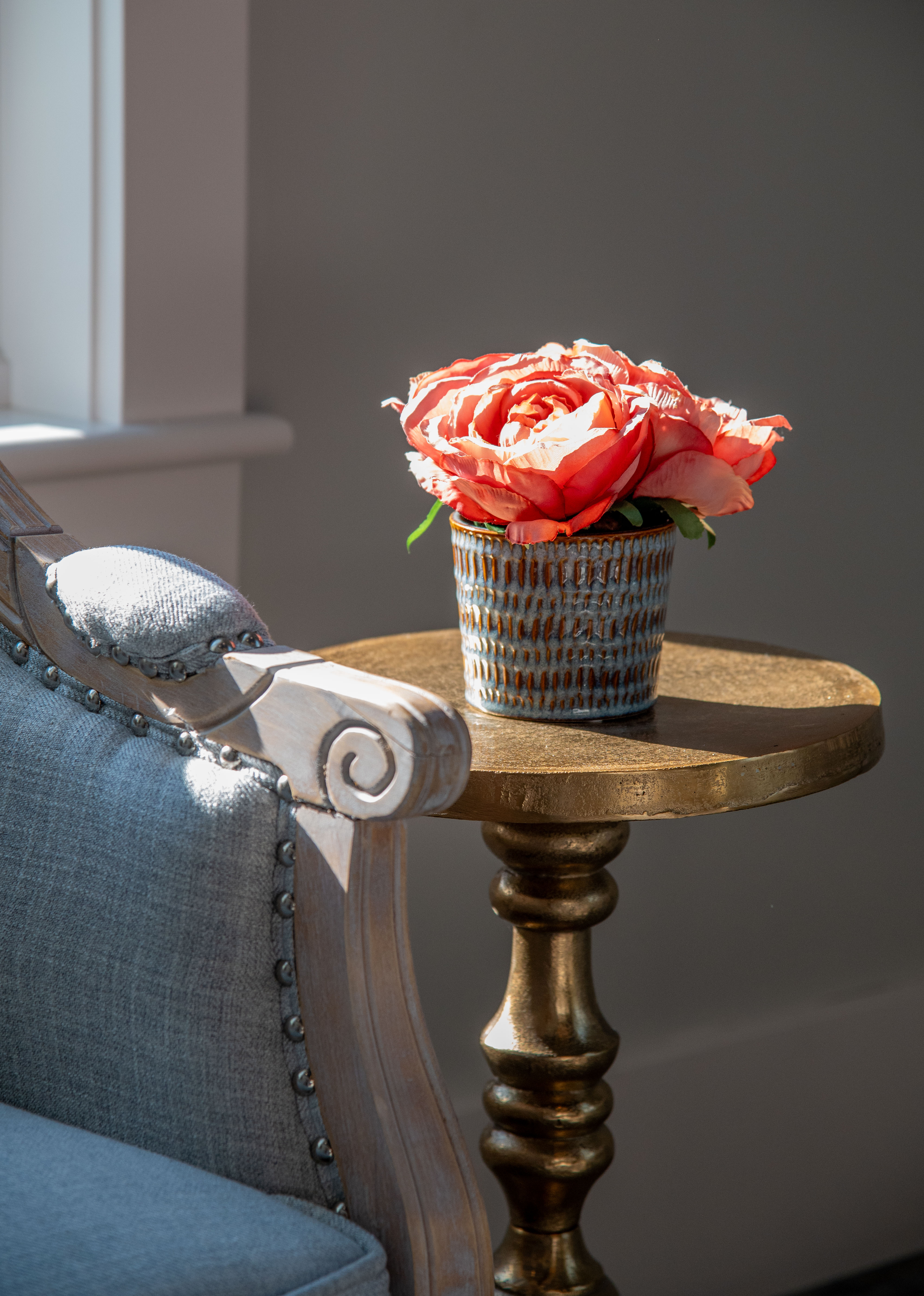 Peach rose in a textured pot on a round golden side table next to a blue upholstered chair.