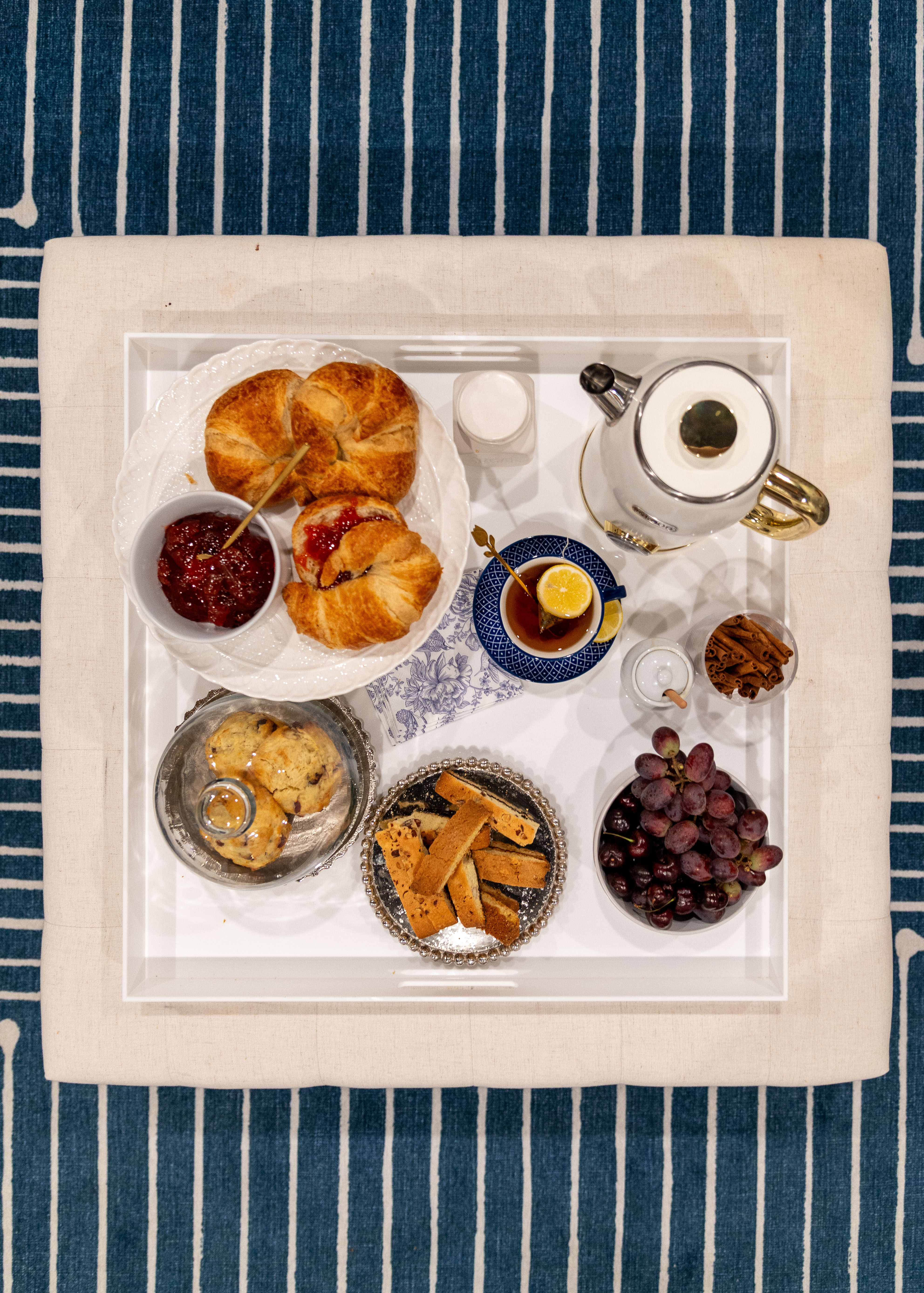 Tray with croissants, jam, cookies, biscotti, tea with lemon, grapes, and cinnamon sticks on a blue patterned carpet.