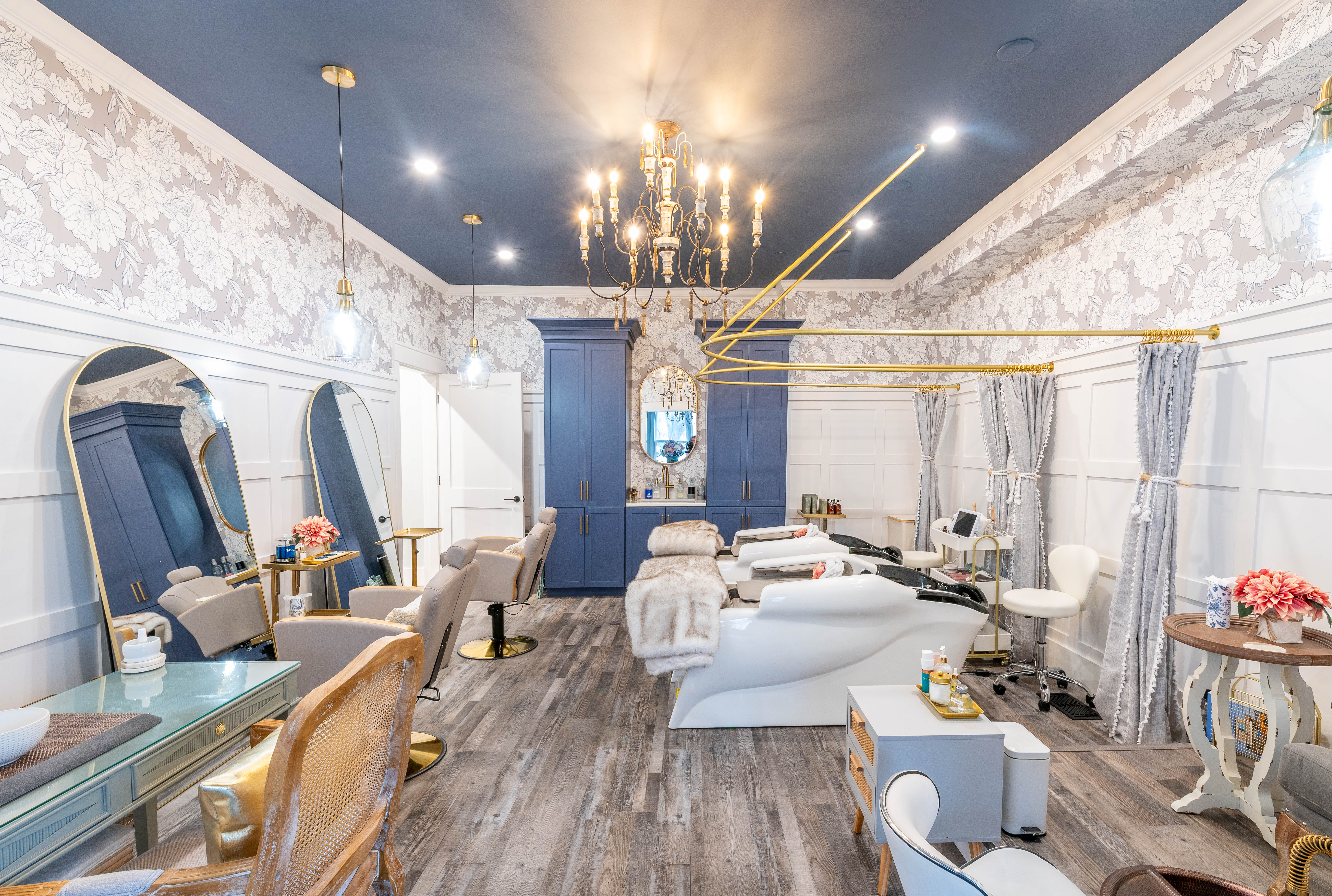 Elegant beauty salon with blue ceiling, chandelier, floral wallpaper, mirrors, wash stations, and salon chairs.