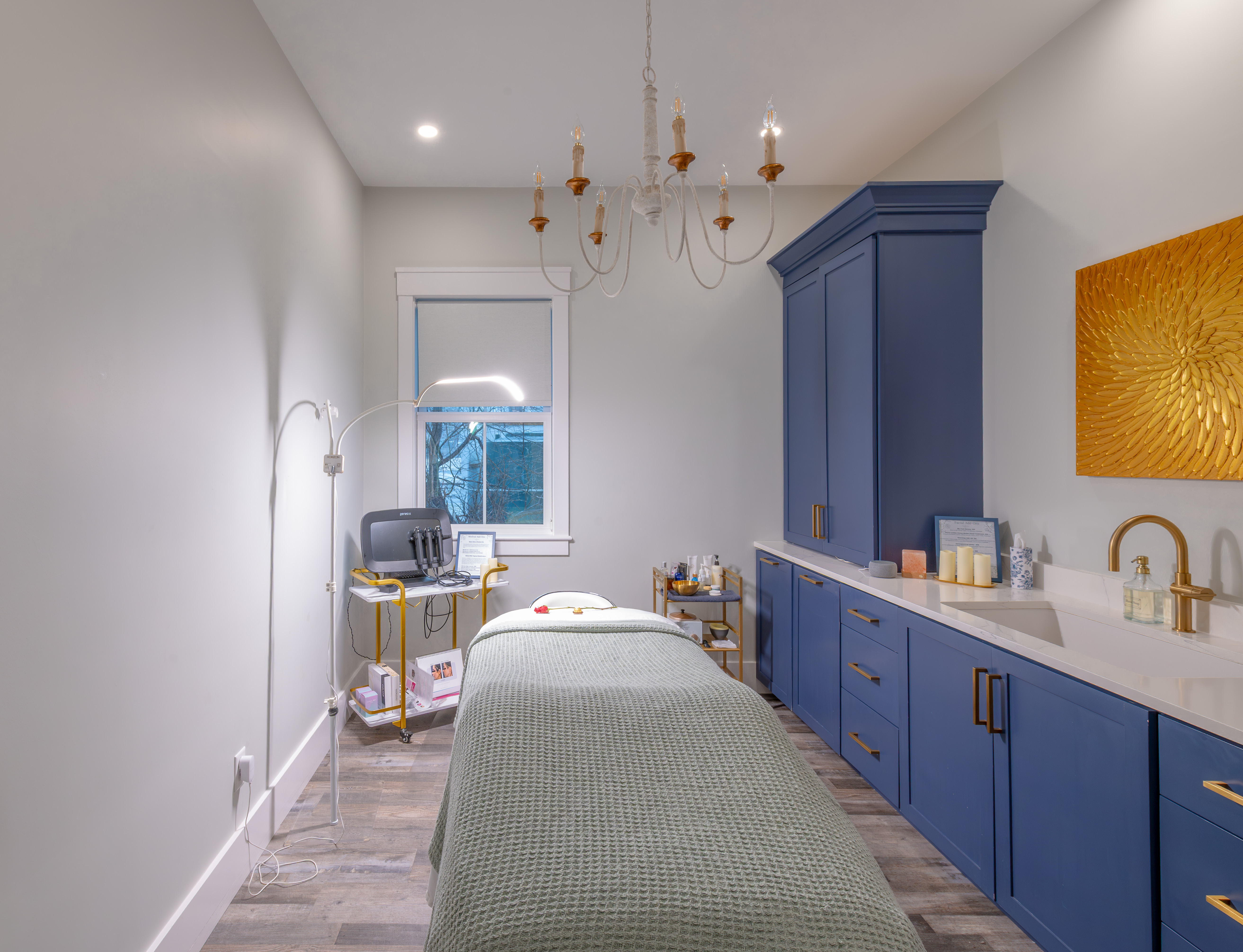 Bright spa room with massage table, blue cabinets, gold faucet, and a golden wall art piece.