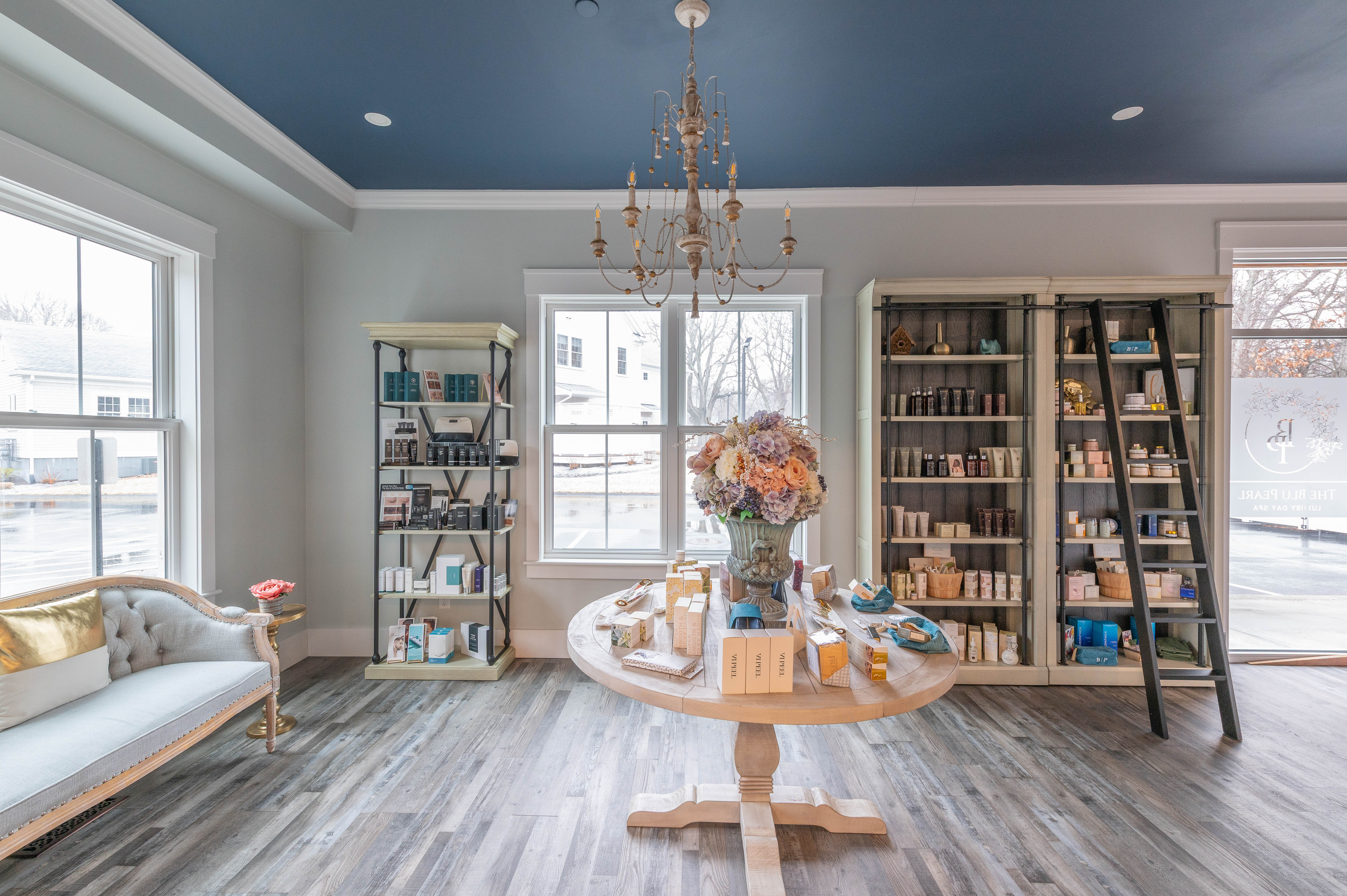 Light-filled spa retail space with shelves, a display table with flowers, and a sofa with gold pillow.