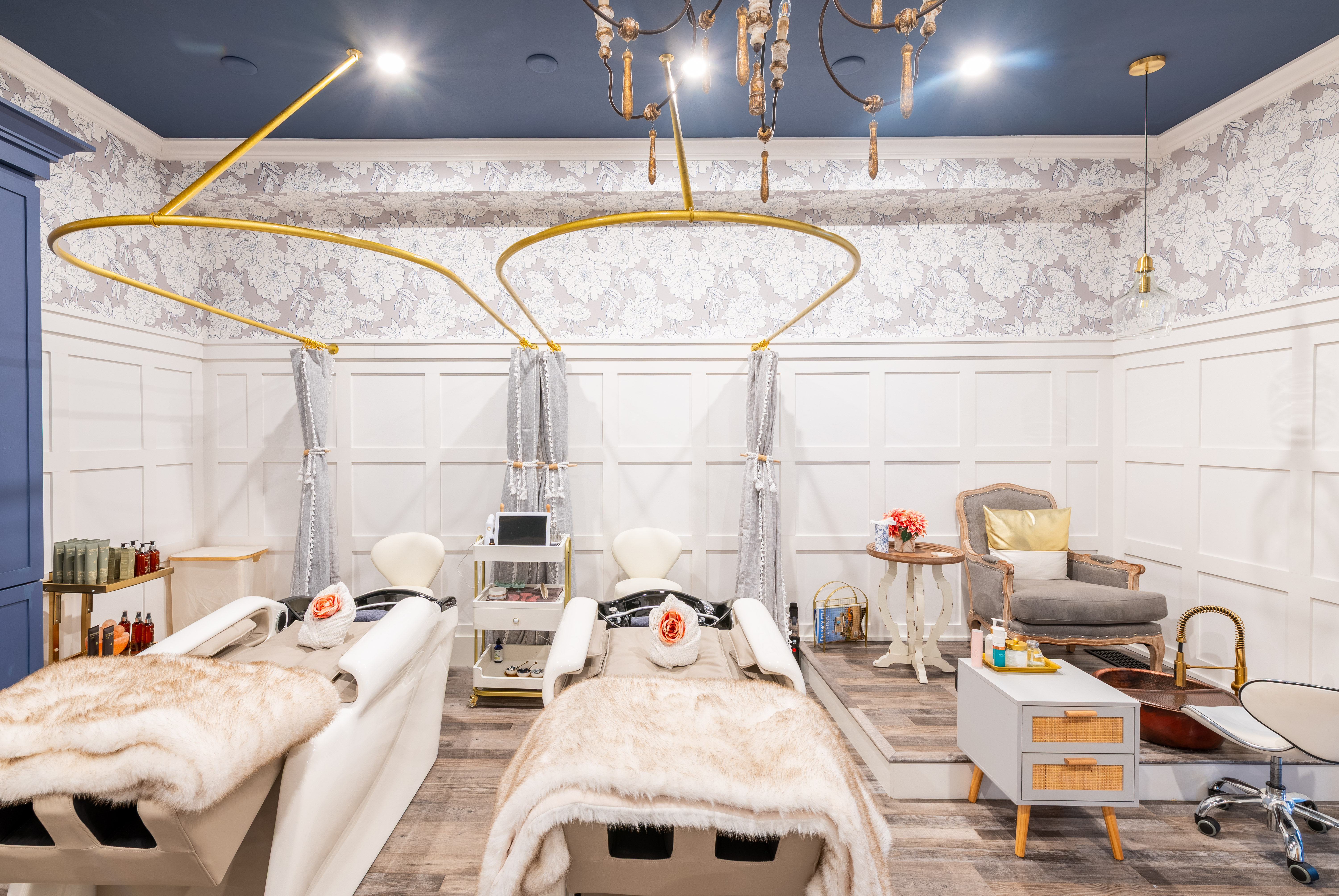 Elegant salon with two white shampoo chairs, floral wallpaper, fur blankets, and cozy seating area.