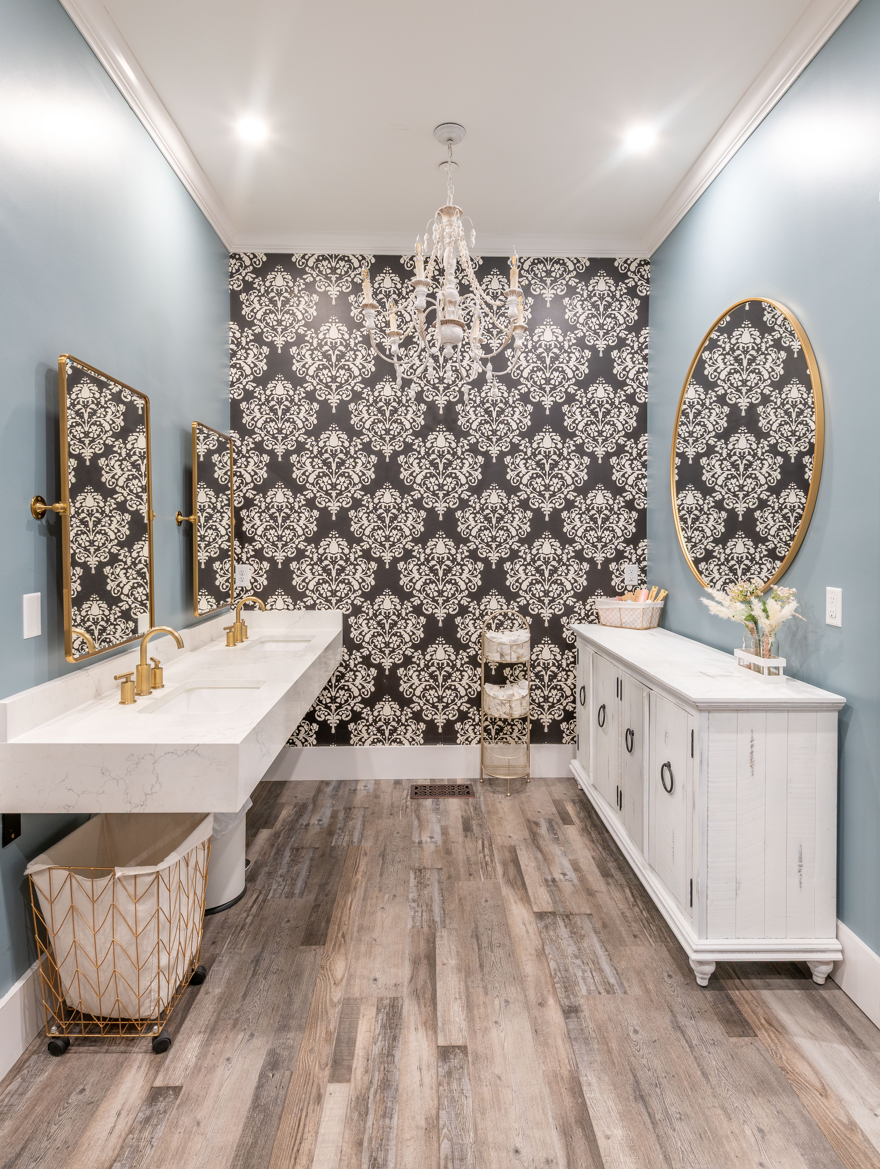 Modern bathroom with double sinks, gold-framed mirrors, patterned black wallpaper, chandelier, and wood flooring.