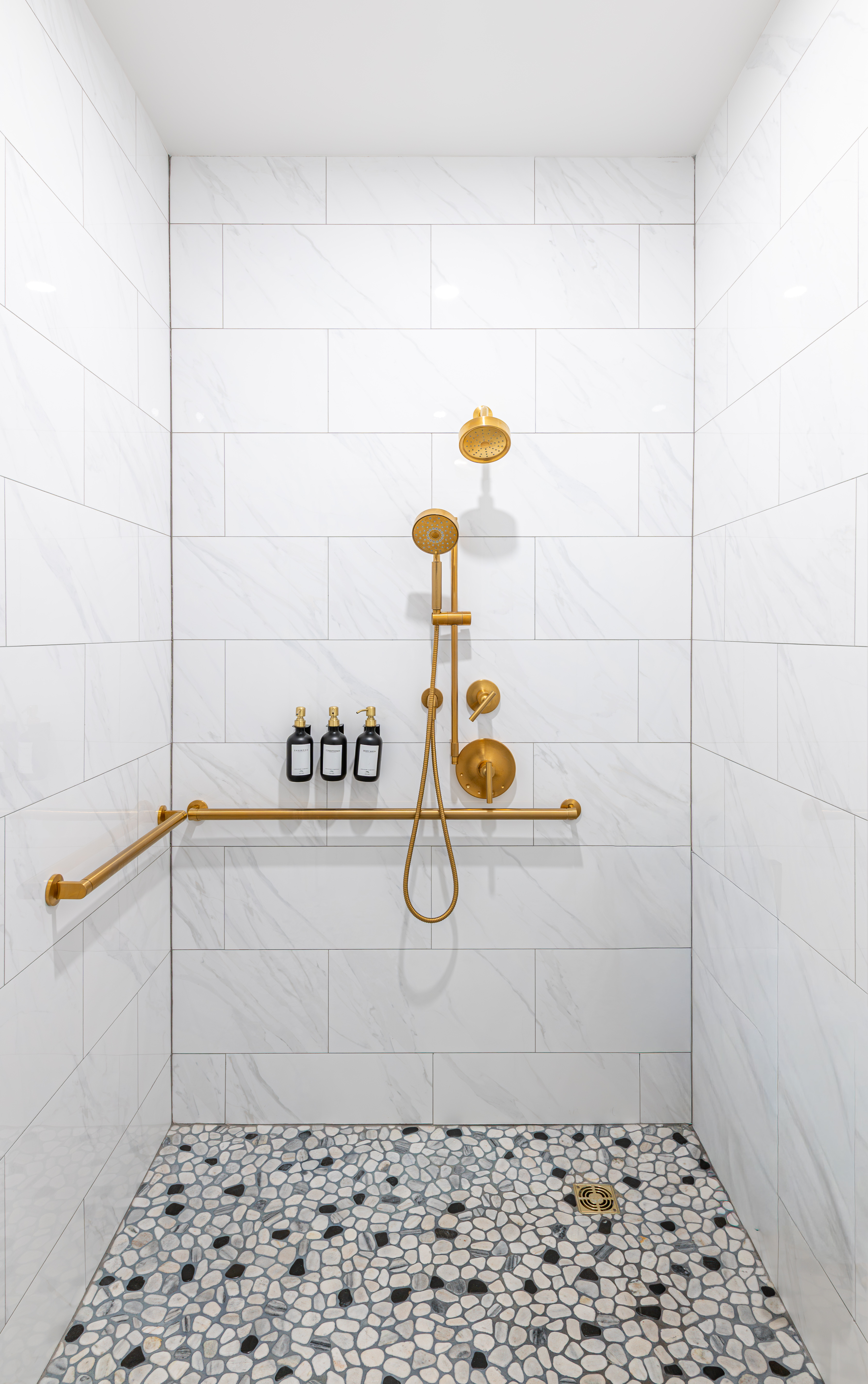 Shower with white marble tiles, pebble stone floor, gold fixtures, and mounted soap dispensers.