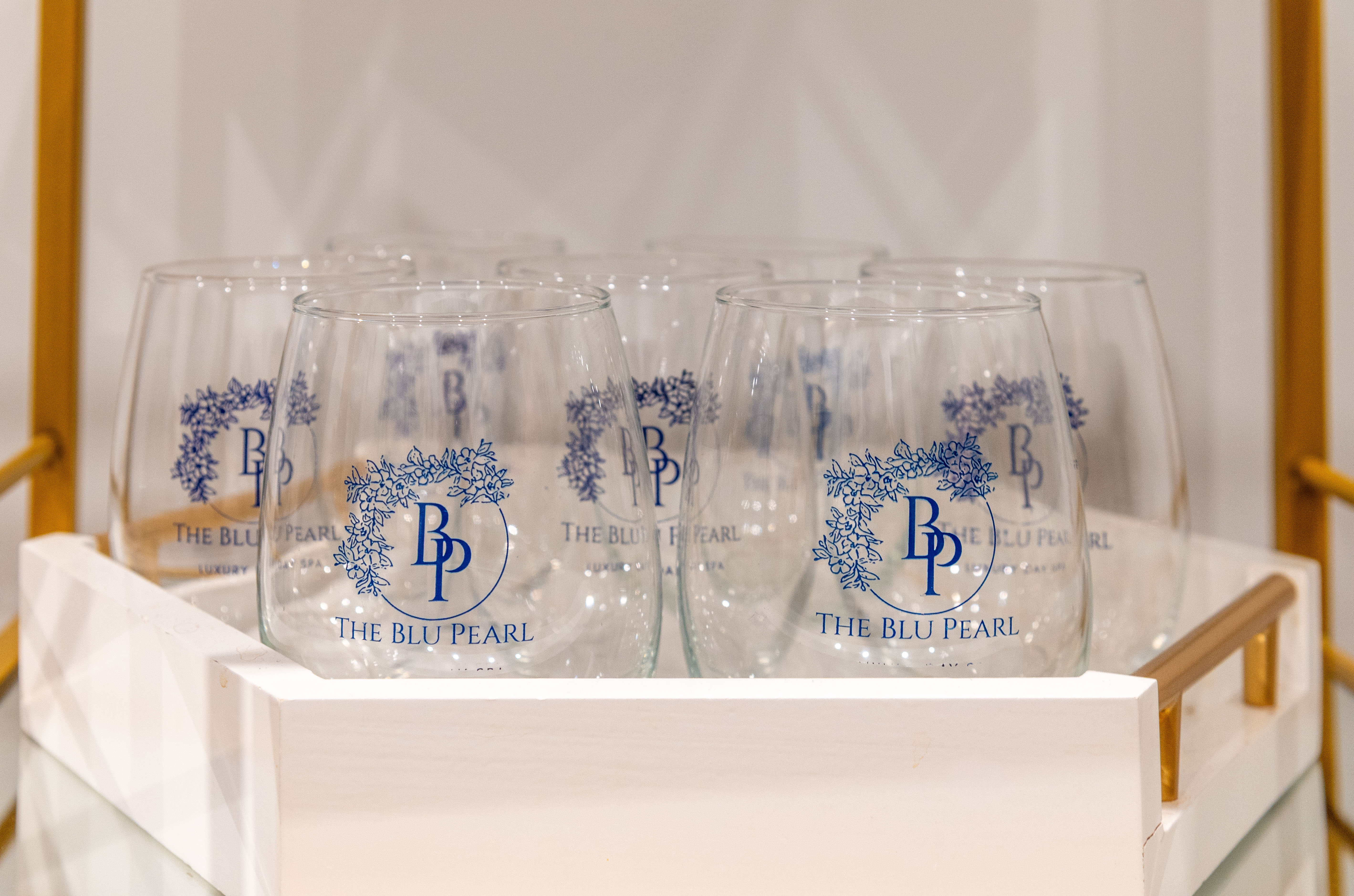 Clear glasses with The Blu Pearl logo arranged in a white tray on a gold and glass cart.