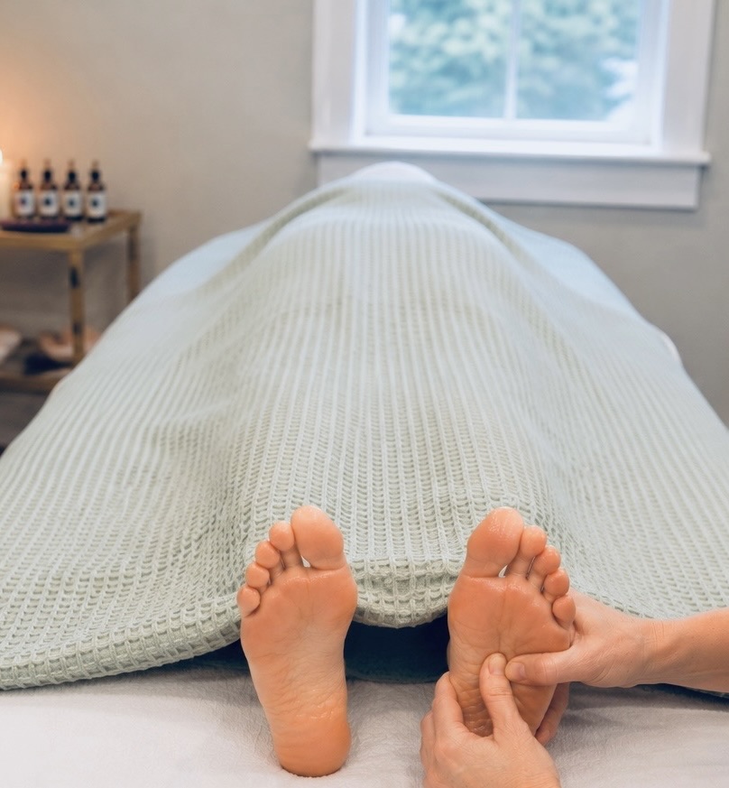 Person covered with blanket receiving a foot massage in a calm room with a window and oils nearby.