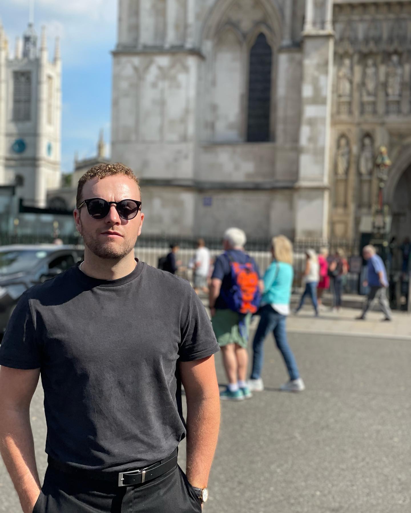 Man with curly hair wearing black sunglasses and a black t-shirt standing in front of a historic stone building on a sunny day.