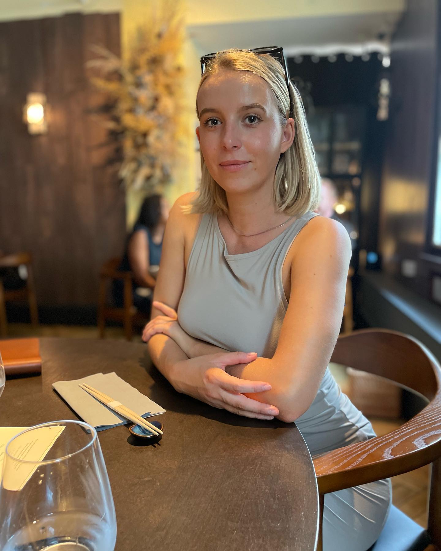 Young woman with blonde hair wearing a sleeveless light gray top sitting at a table with chopsticks and a napkin in a softly lit restaurant.