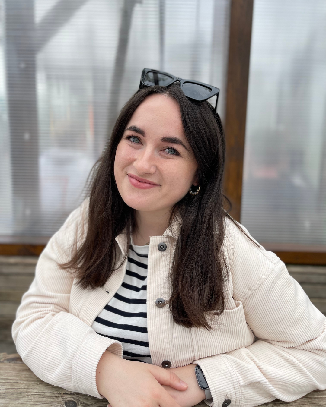 Smiling woman with long dark hair wearing a cream corduroy jacket and black-striped shirt, resting her arms on a wooden surface.