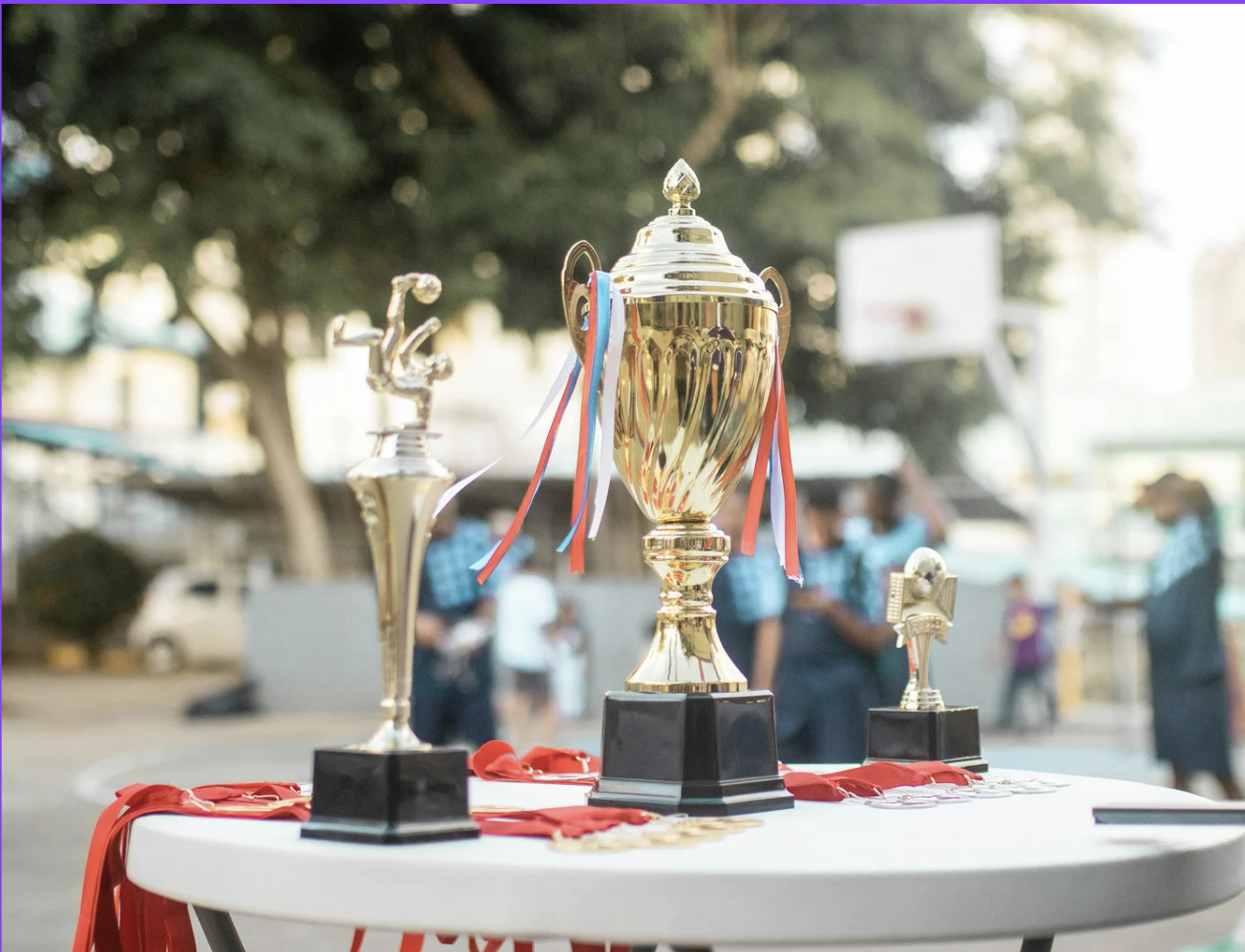 Three trophies with red and blue ribbons displayed on a white table outdoors with blurred people and trees in the background.