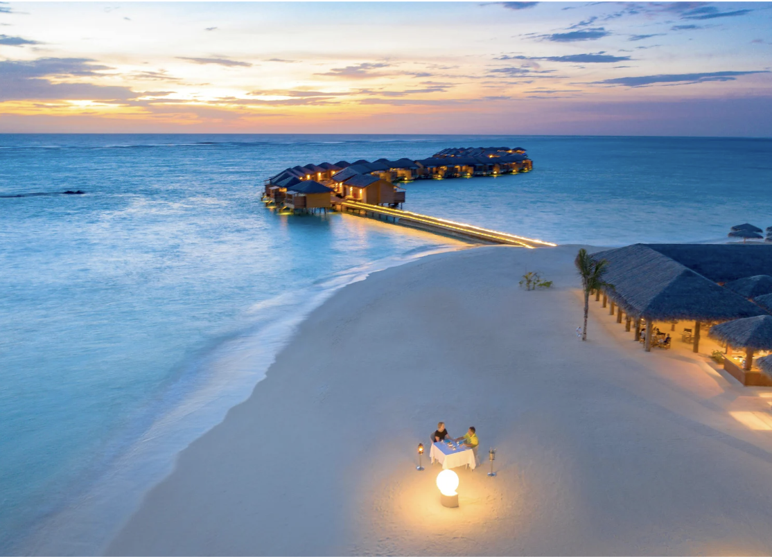 Couple dining at a candlelit table on a sandy beach with overwater bungalows and a sunset-lit sky in the background.