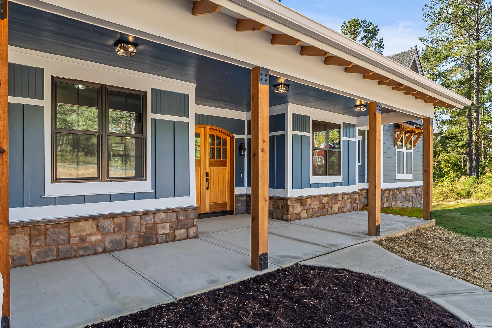 Front porch of a modern house featuring stone siding, wooden columns, blue-gray walls, and a wooden front door.