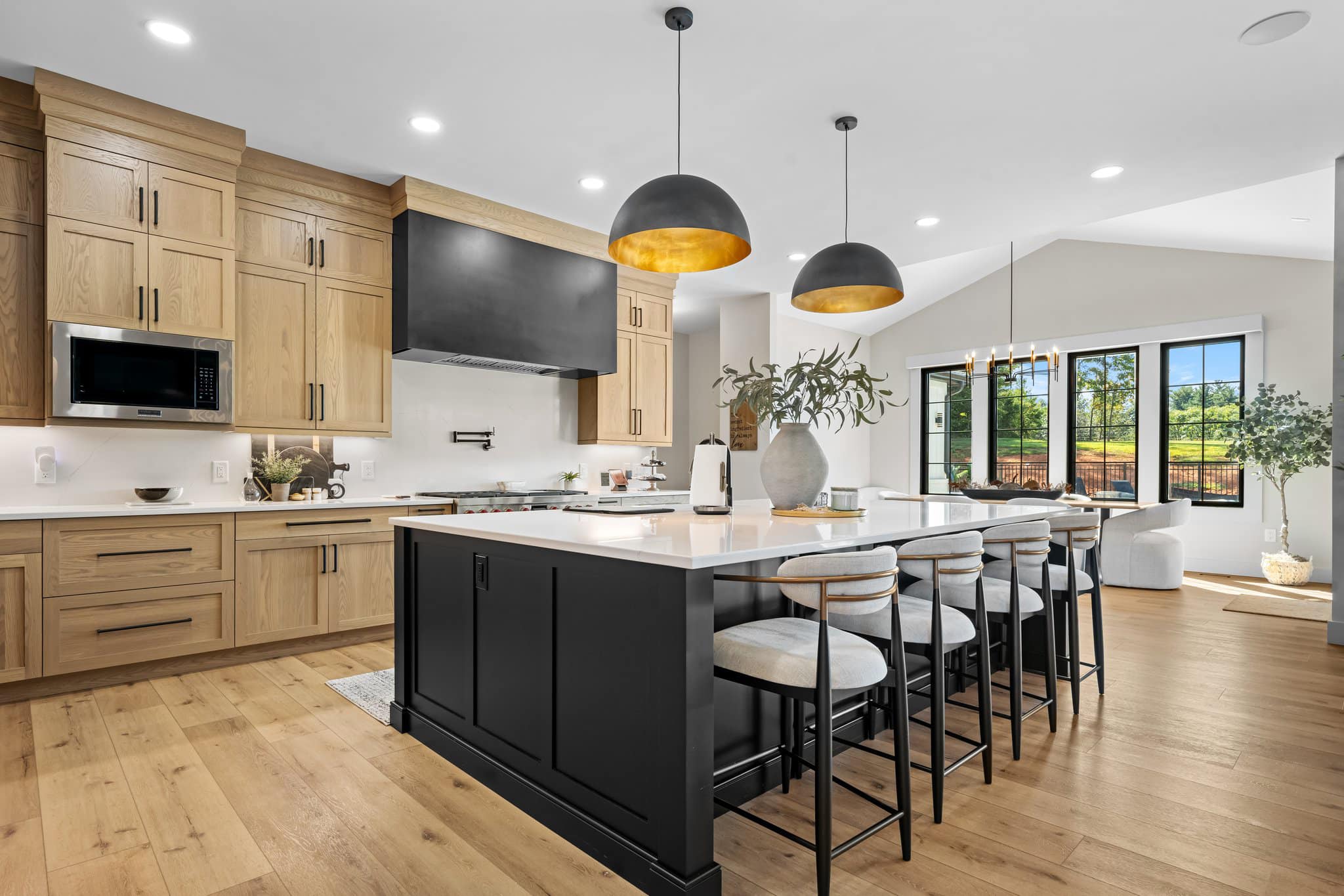 Modern kitchen with light wood cabinets, large black island with white countertop, pendant lights, and bar stools, adjacent to a dining area with large windows.