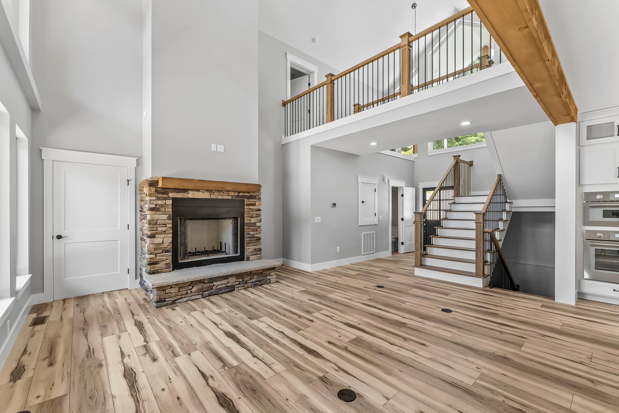 Spacious modern living room with light wood flooring, stone fireplace, and wooden staircase leading to a loft area.