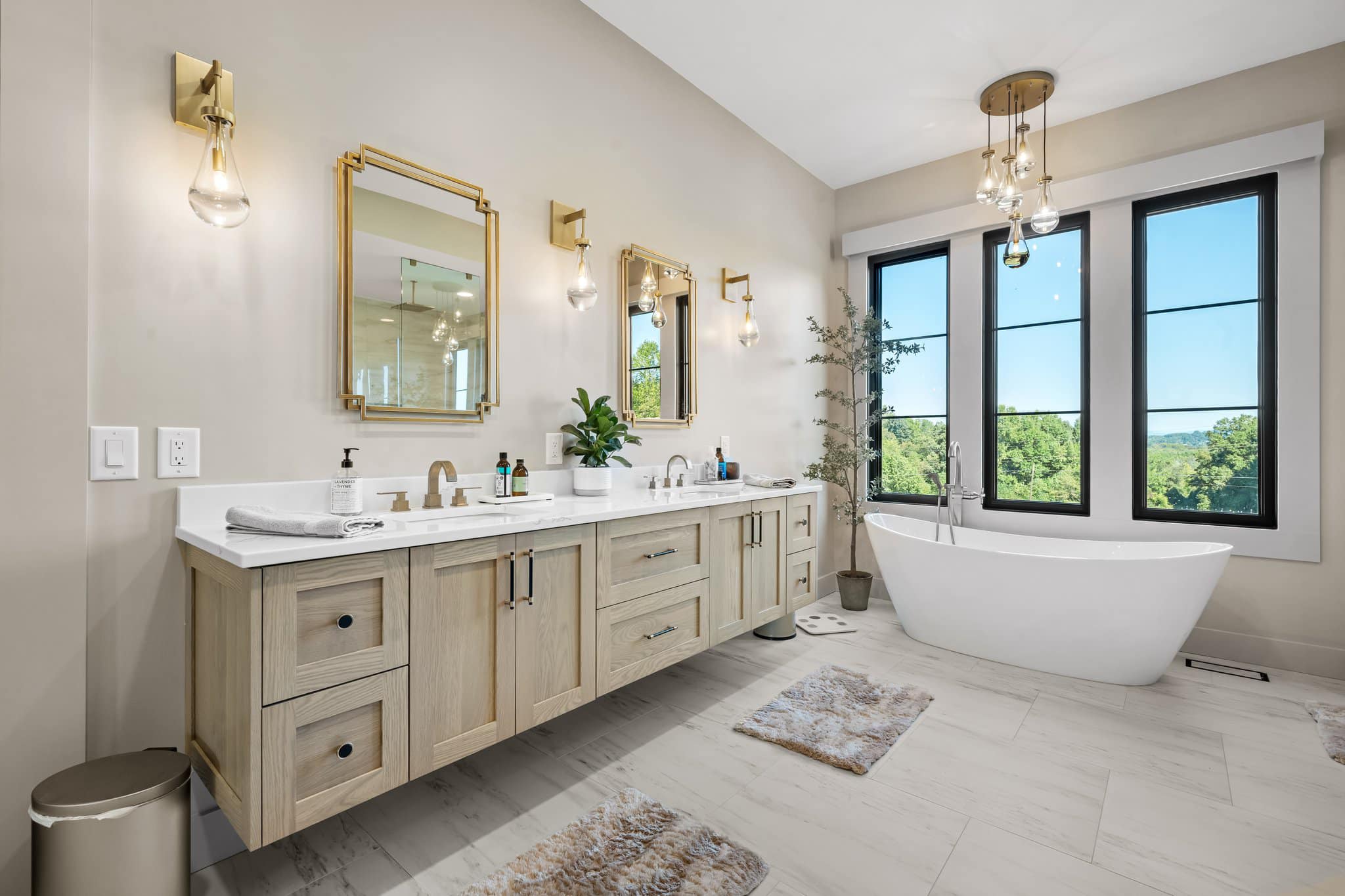 Modern bathroom with double vanity, gold-framed mirrors, pendant lights, freestanding bathtub, and three large windows showing greenery outside.