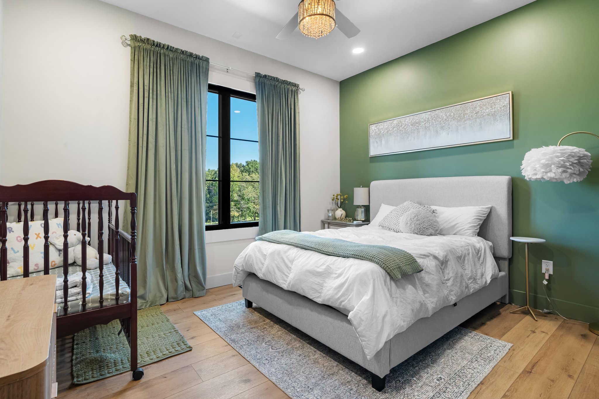 Modern bedroom with a green accent wall, gray upholstered bed, green curtains, wooden crib, and a ceiling fan with a chandelier light.