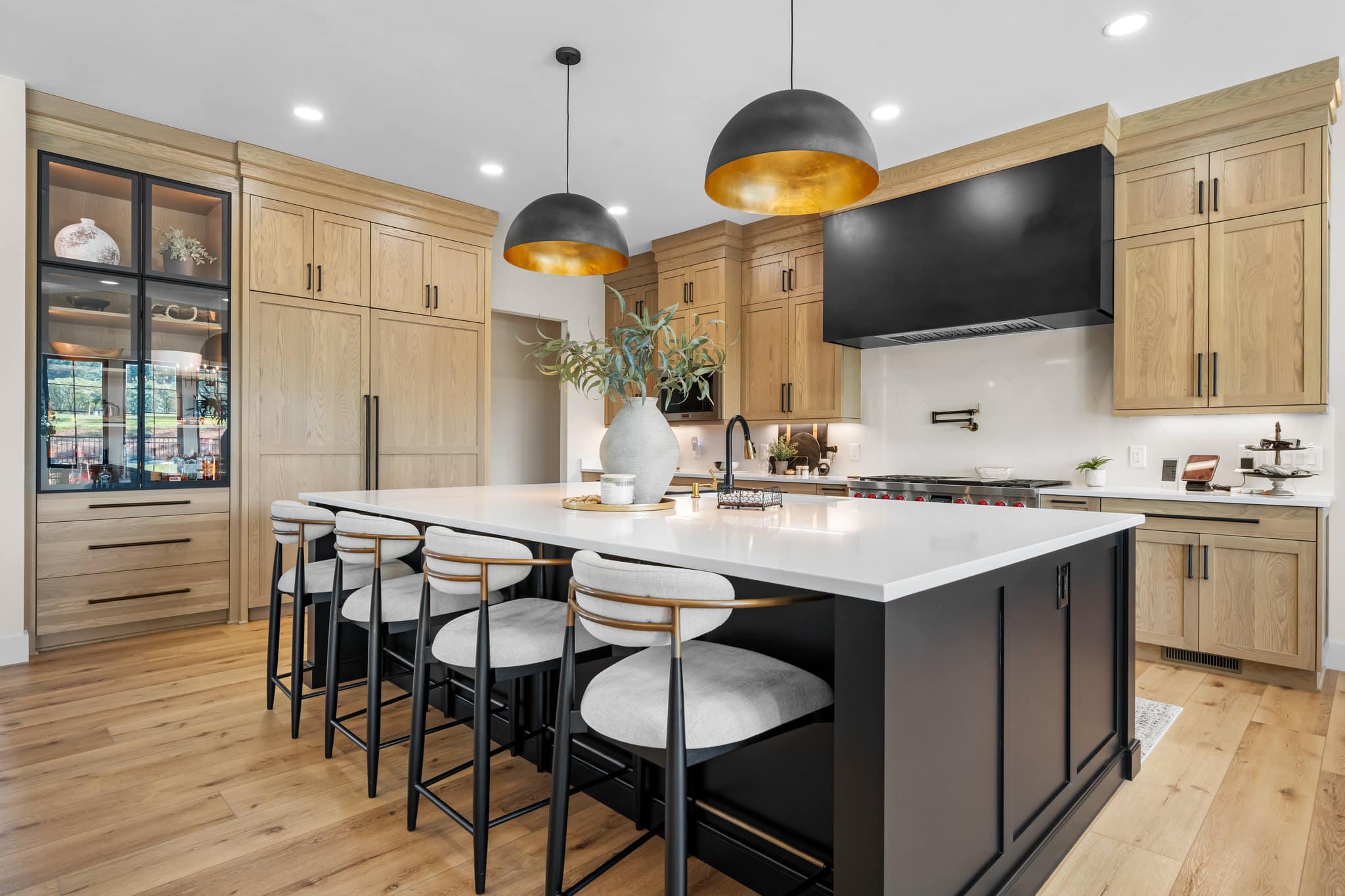 Modern kitchen with large white island, black base, five cushioned bar stools, wooden cabinets, and two black pendant lights with gold interiors.