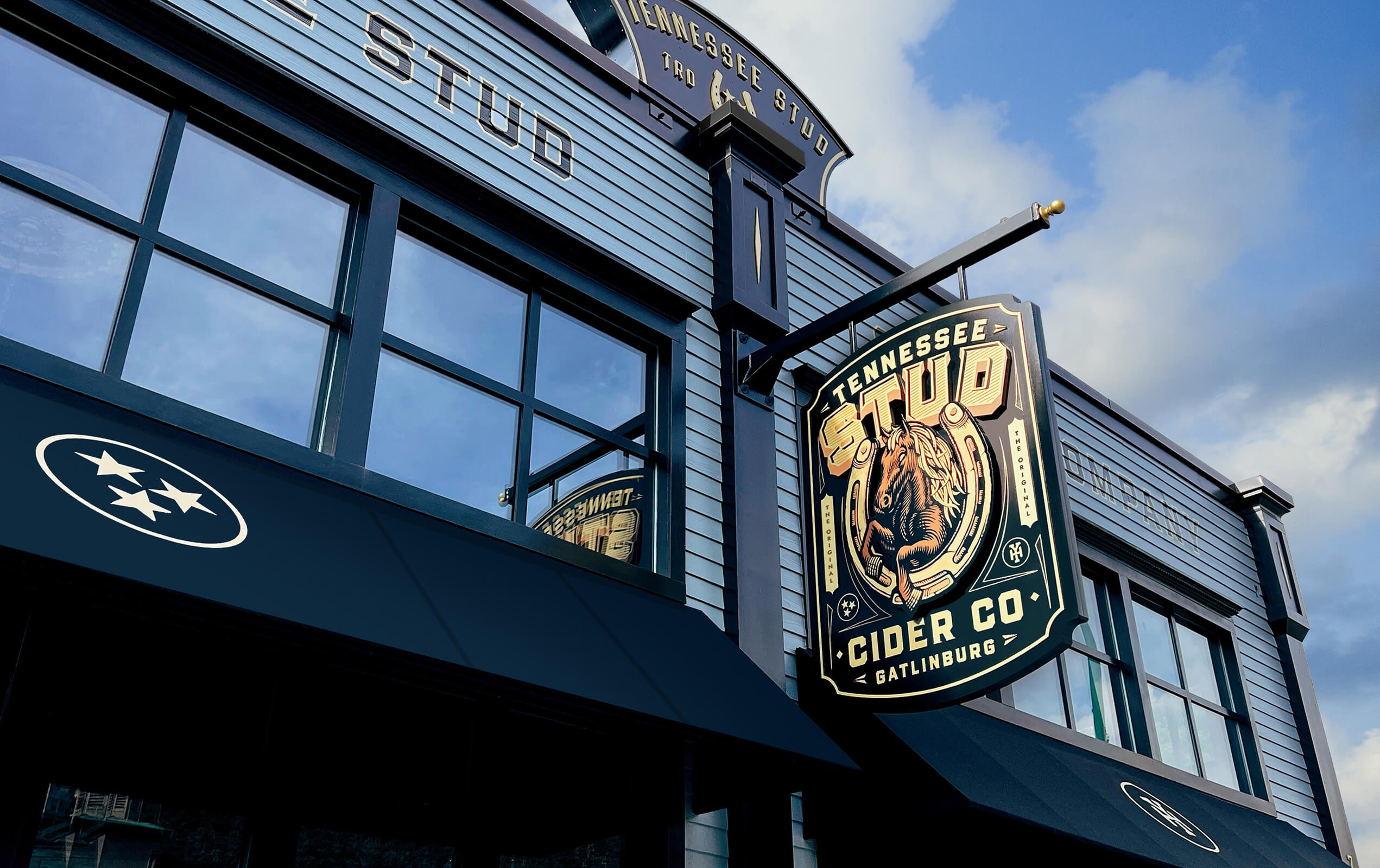 Exterior of a building with blue siding and large windows reflecting the sky, featuring a hanging sign that reads 'Tennessee Stud Cider Co Gatlinburg' with an illustrated horse inside a horseshoe, and black awnings with a white three-star emblem.