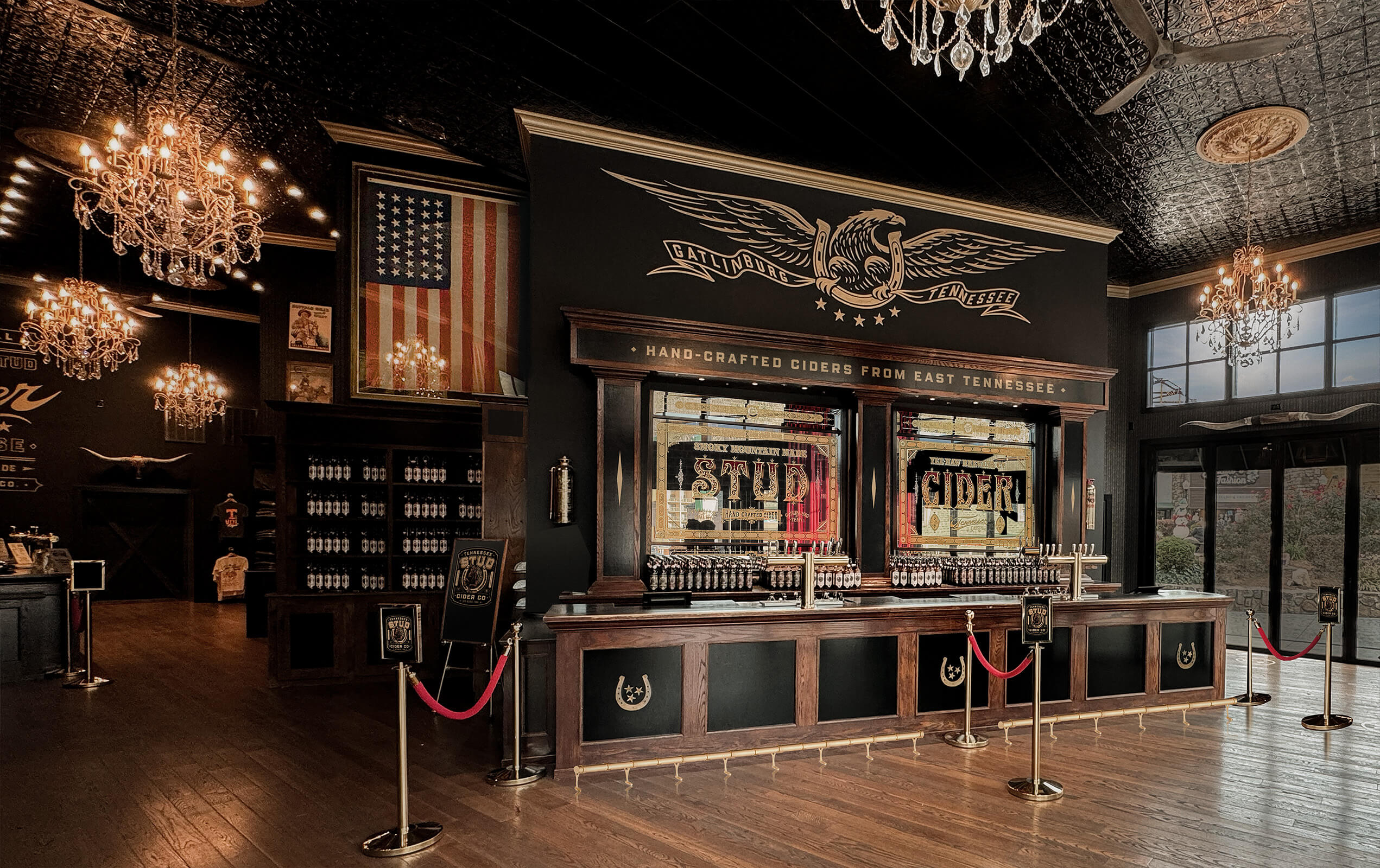 Elegant cider bar interior with dark wood counters and stained glass windows reading 'Stud Cider', American flag on the wall, chandeliers hanging from an ornate black ceiling, and product shelves with cider bottles.