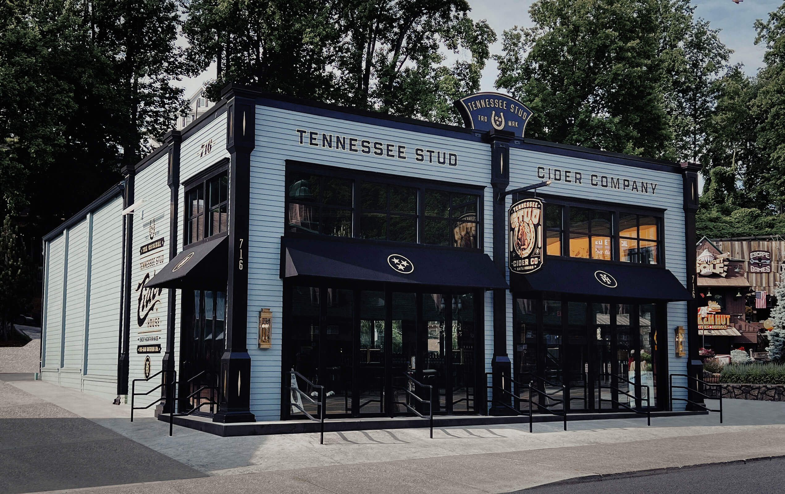 A blue two-story building with black trim housing Tennessee Stud Cider Company, with large windows, black awnings, and signage, surrounded by trees and other buildings.