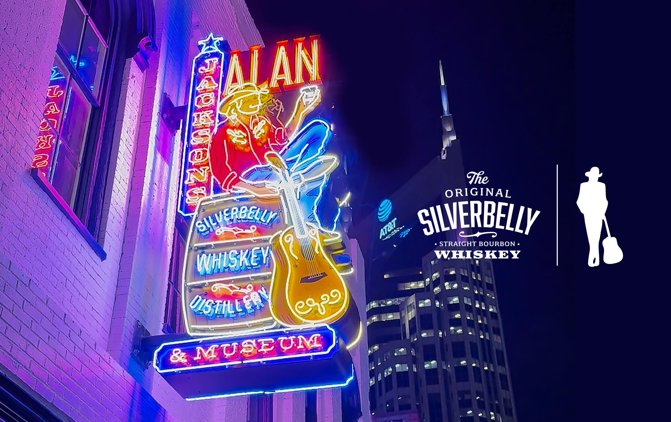 Bright neon sign on a building exterior at night reading 'JACKSON'S ALAN SILVERBELLY WHISKEY DISTILLERY & MUSEUM' with an illustration of a seated cowboy playing guitar. City skyscraper with AT&T logo is visible in the dark sky background. The right side displays a logo for The Original Silverbelly Straight Bourbon Whiskey alongside a silhouette of a person with a hat and guitar case.