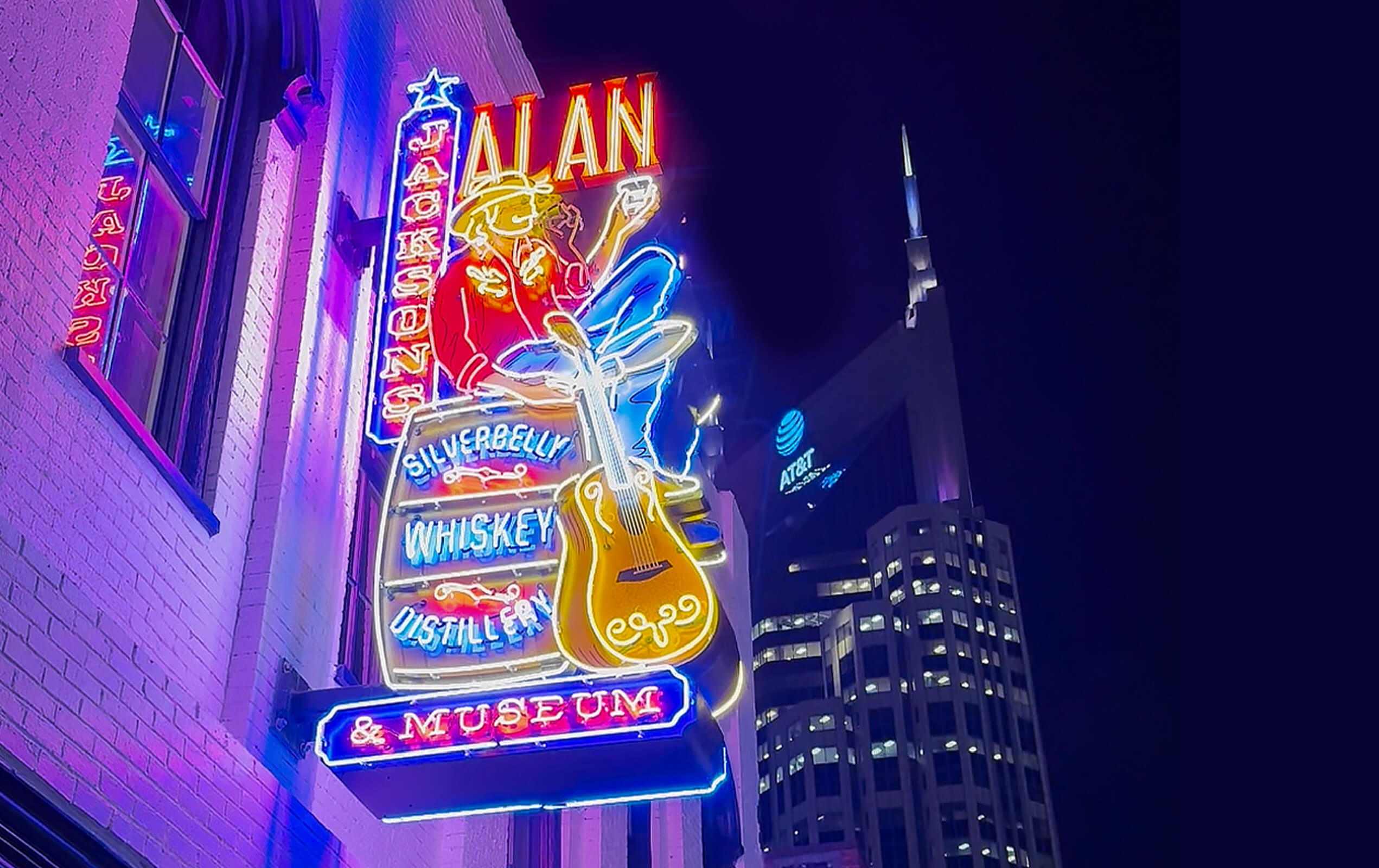 Neon sign for Jack Daniel's Silver Bell Whiskey Distillery & Museum featuring a cowboy playing a guitar, mounted on a building at night with a lit AT&T skyscraper in the background.