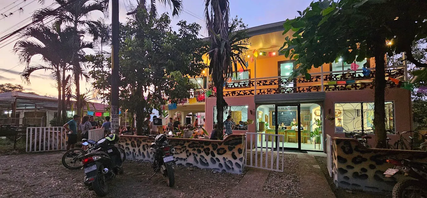 Evening view of a lively café with outdoor seating, colorful decorations, and motorcycles parked outside.