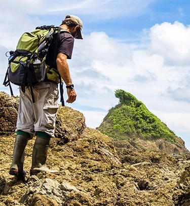 Man with backpack and boots hiking on rocky terrain with a green hill and cloudy sky in background.
