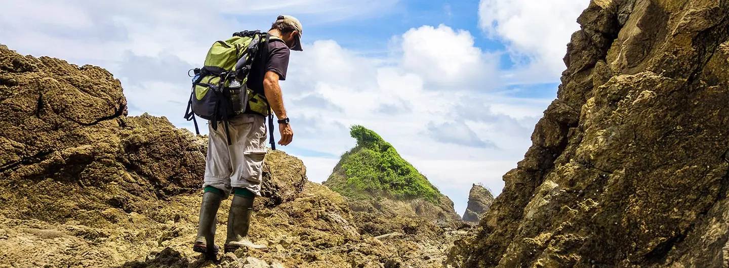 Person wearing backpack and boots hiking over rocky terrain under a partly cloudy sky.
